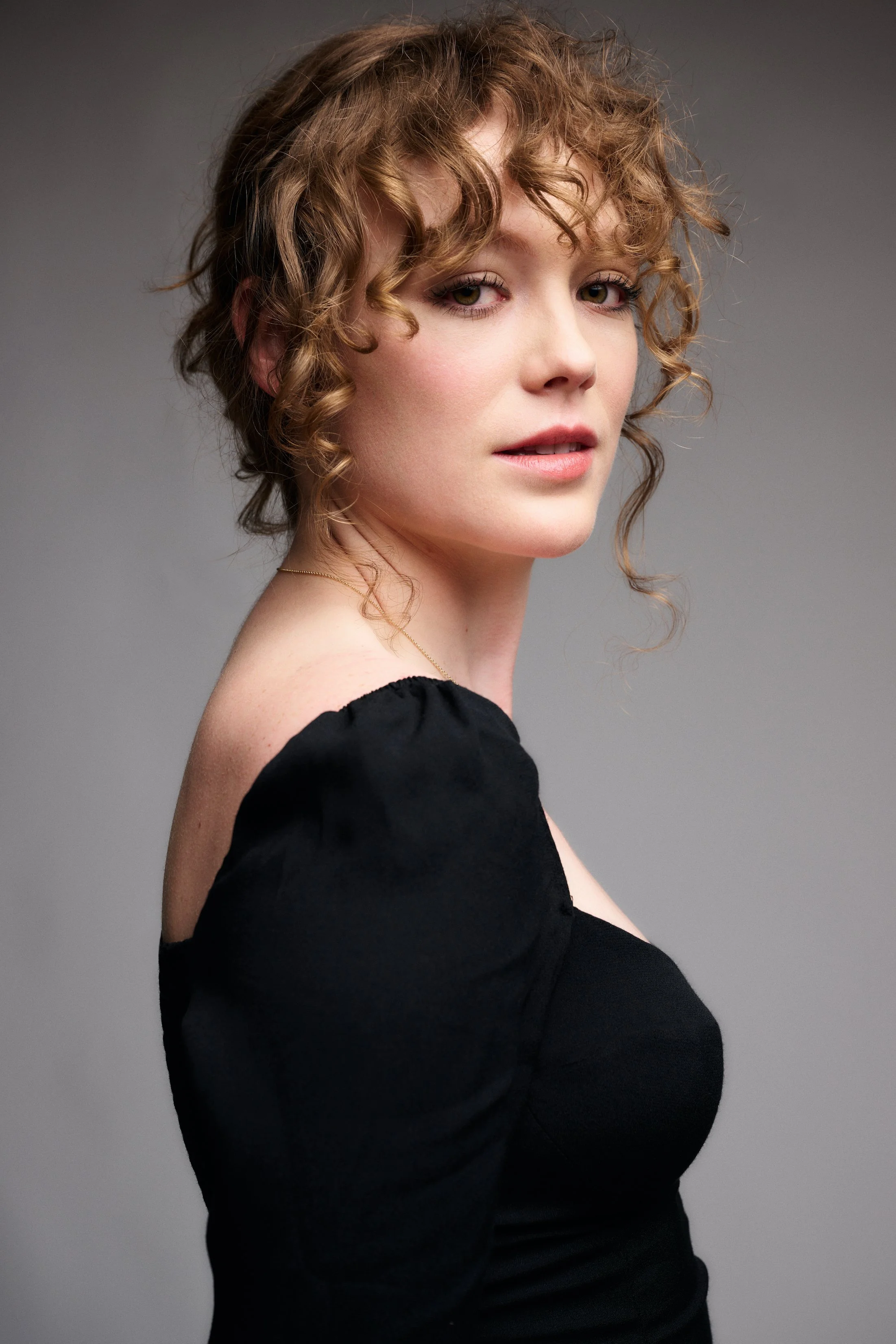 Close-up portrait of Holly Hinchliffe with curly light brown hair, wearing a black top, against a gray background.