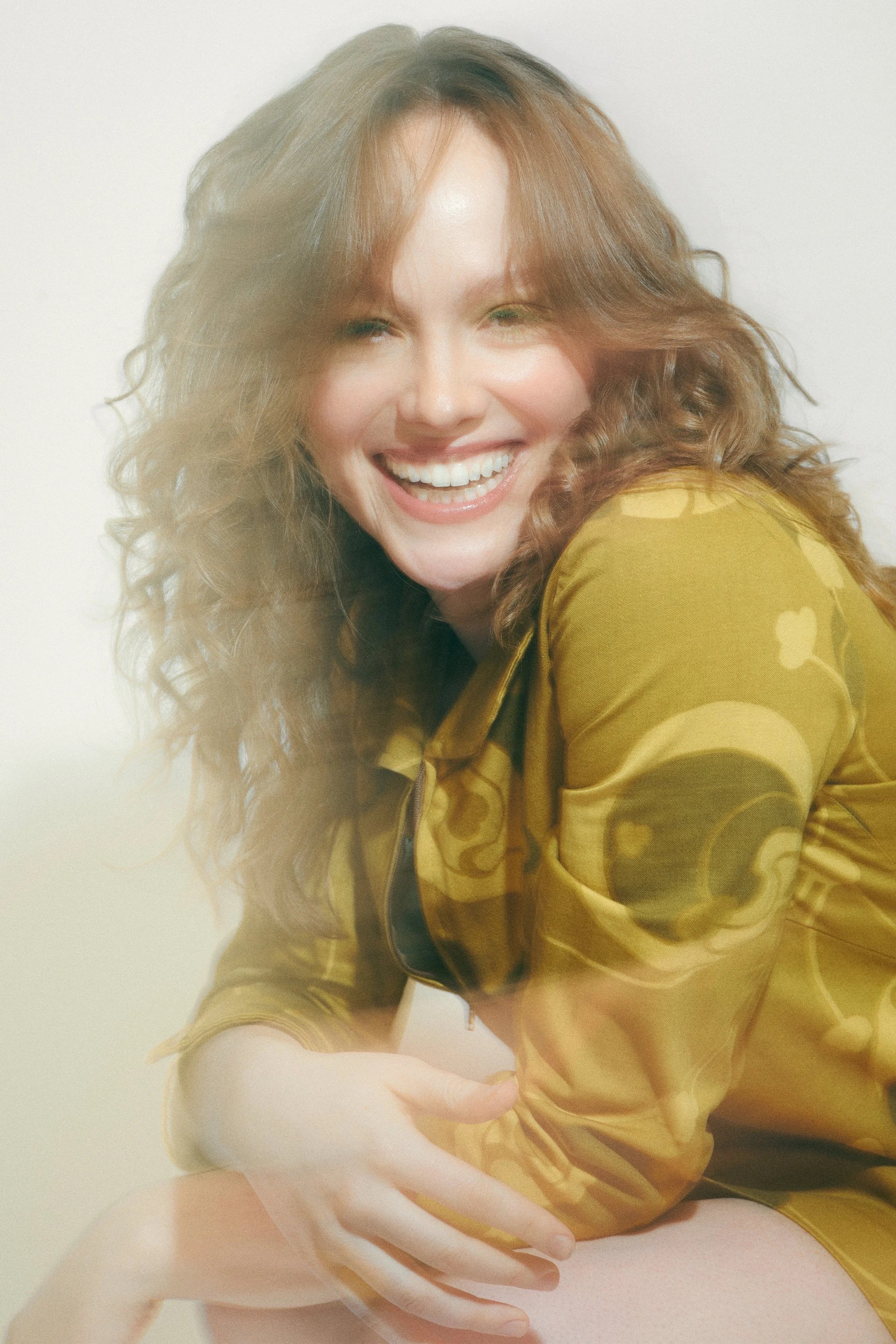 Holly Hinchliffe with curly auburn hair smiling brightly, wearing a gold shirt with a swirling pattern, posing against a neutral light background.