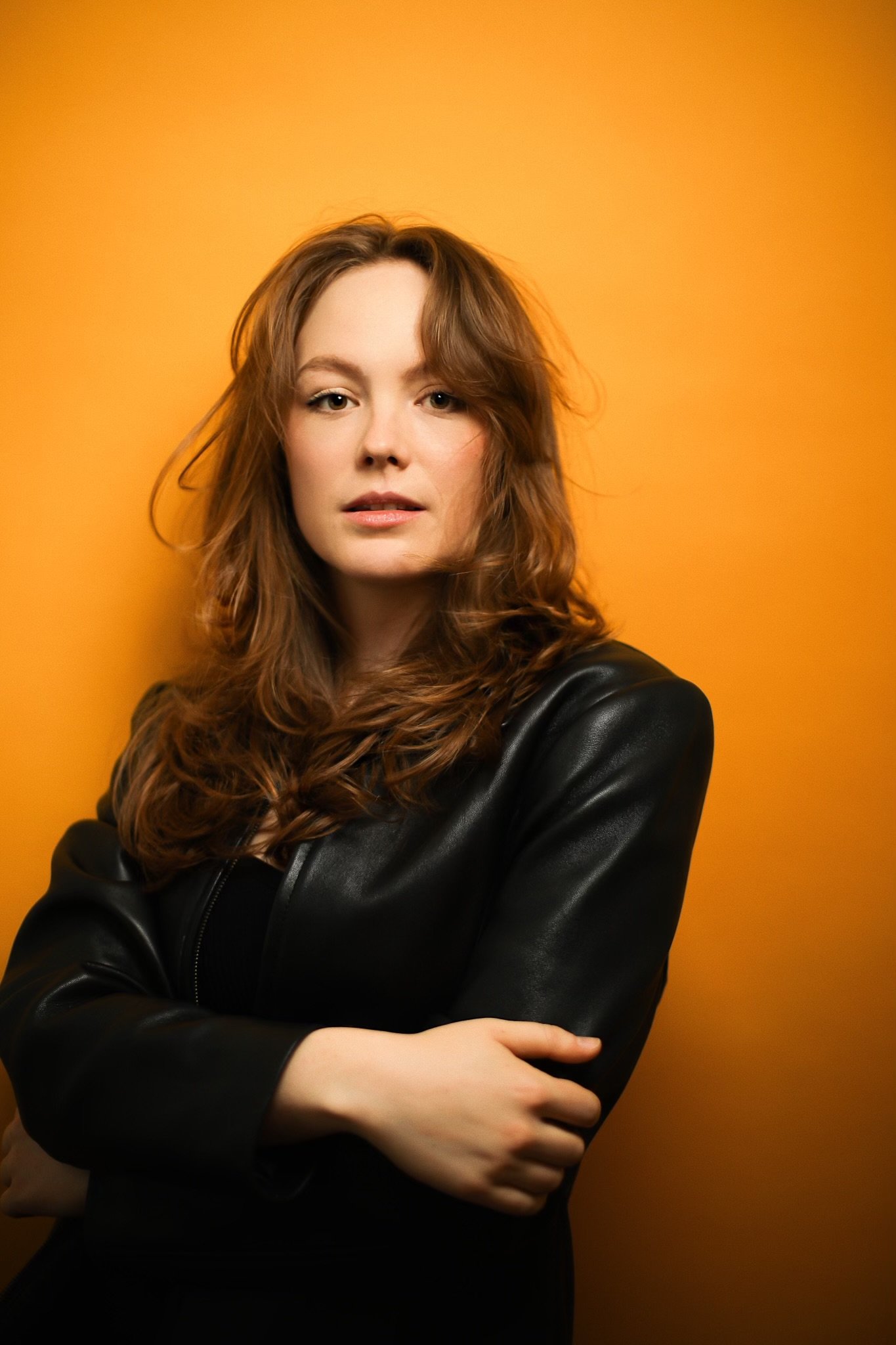 Holly Hinchliffe with wavy red hair in a leather jacket standing against an orange background.