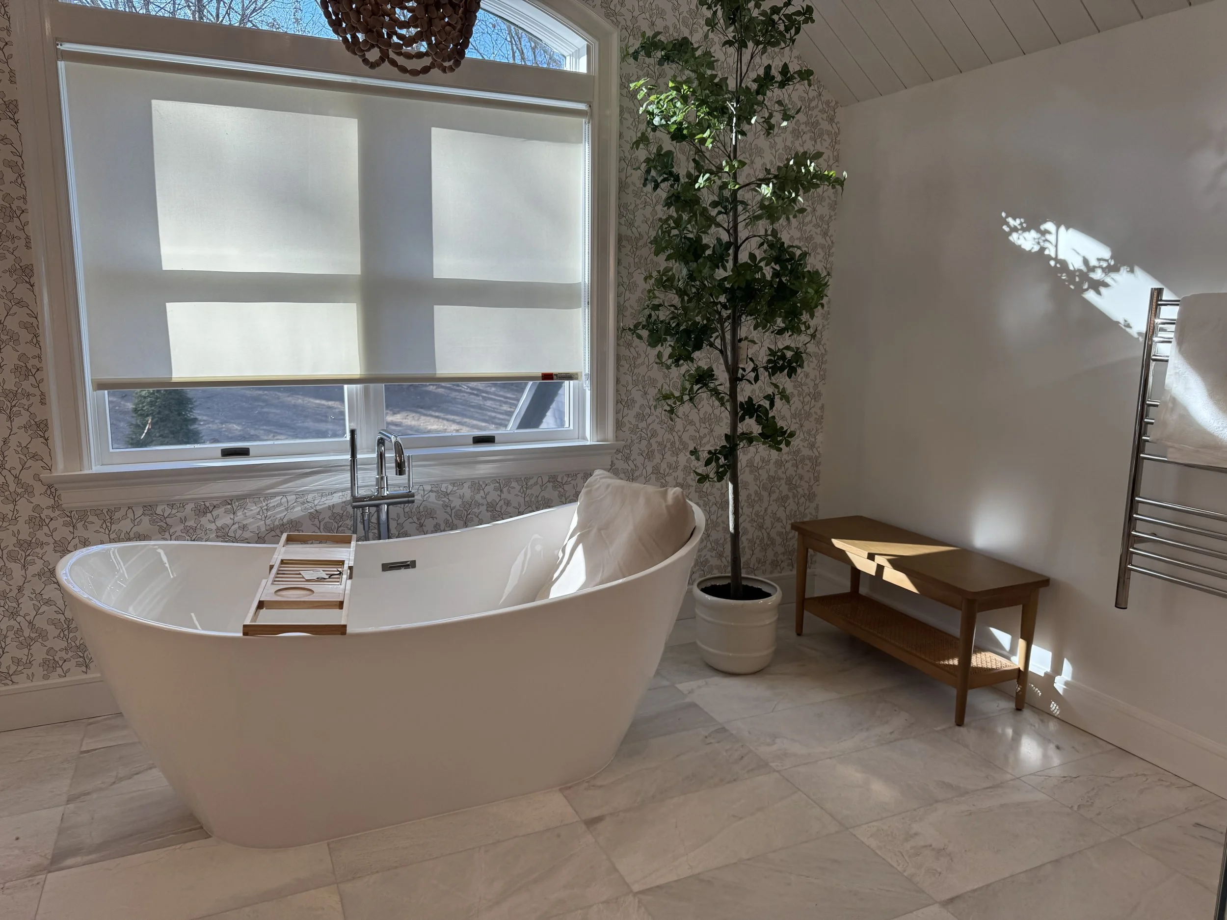Bathroom with a white freestanding bathtub, a faucet, a window with frosted blinds, a potted tall plant, a wooden bench, and a towel warmer on the wall.