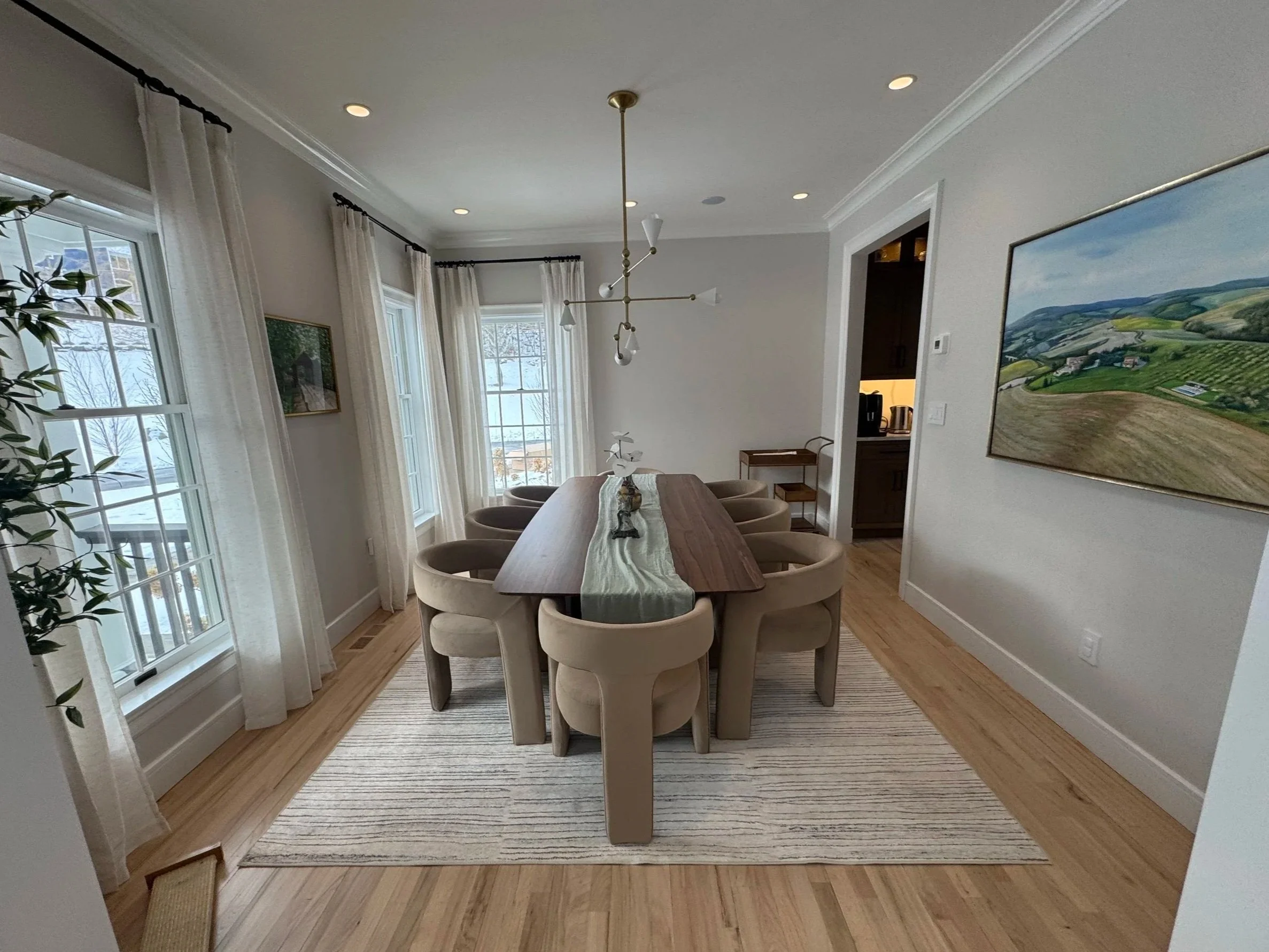 A dining room with a long wooden table, surrounded by beige upholstered chairs, white walls, and large windows with white curtains. There is a modern chandelier overhead and artwork on the walls. A kitchen is visible through an open doorway.
