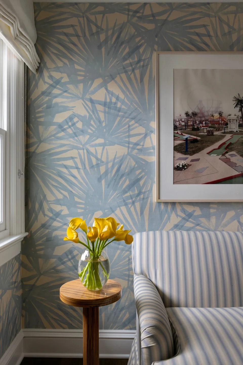 A living room corner with a blue and cream striped armchair, a small wooden side table with a glass vase of yellow calla lilies, a wall with blue palm leaf patterned wallpaper, a framed picture of an amusement park, and a window with white curtains.