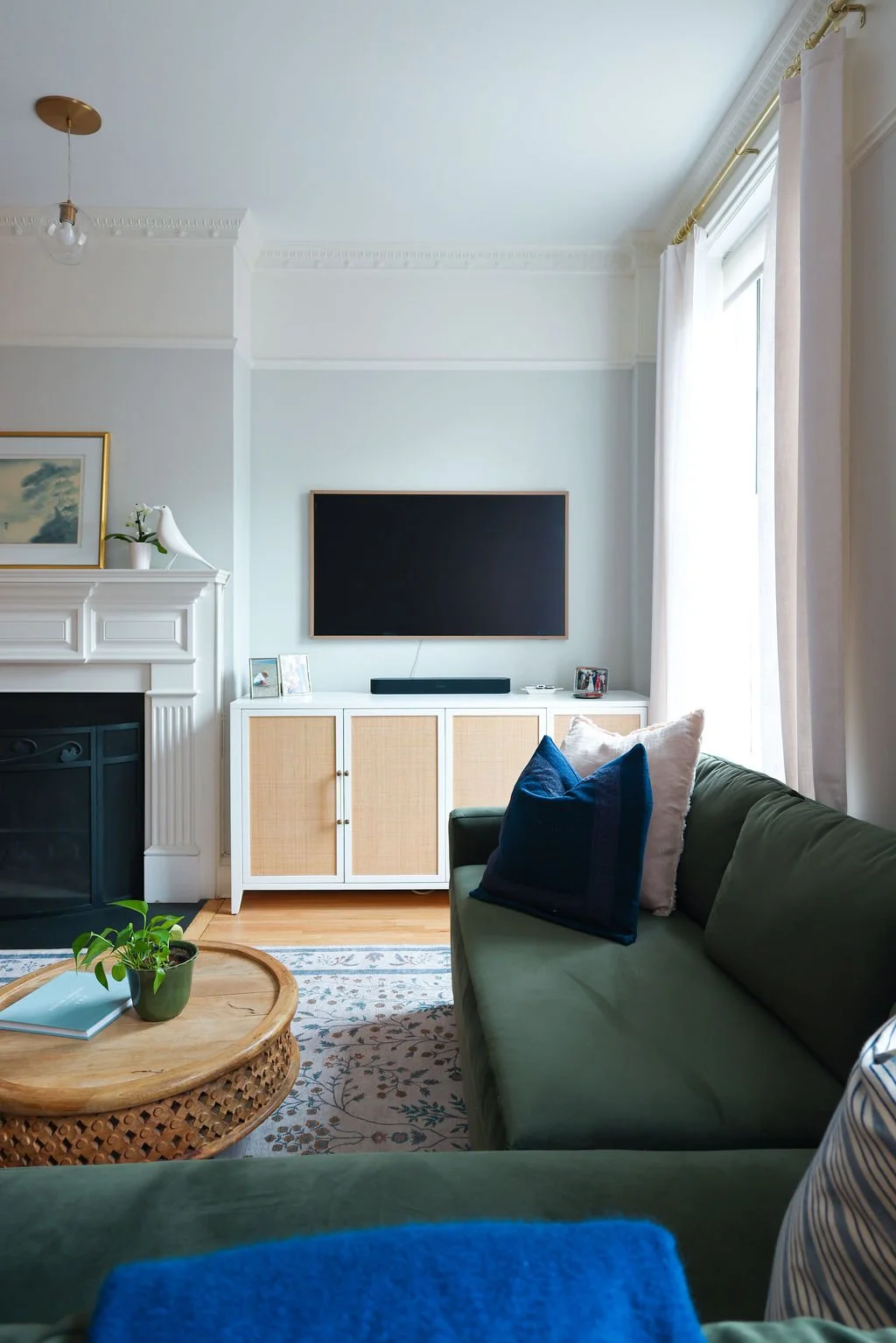 Living room with white fireplace, green sofa with pillows, light wood and white media console, mounted flat-screen TV, window with curtains, and a round wooden coffee table with a plant and books.