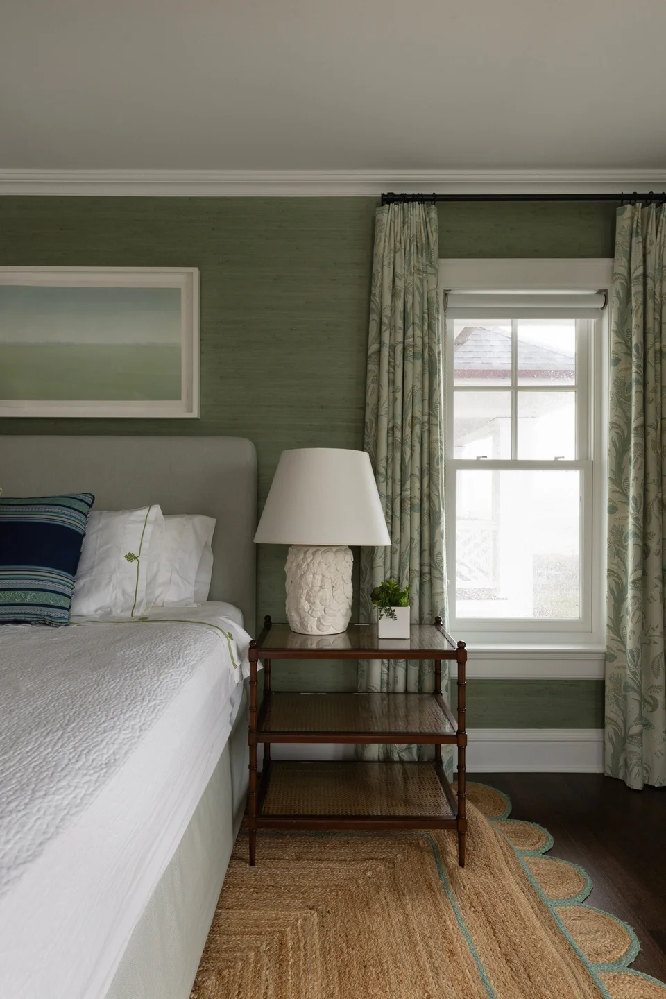 A bedroom with a bed, a green textured wall, a window with open curtains showing a neighboring house roof, a nightstand with a large white seafood-themed lamp, a small potted plant, and an abstract painting on the wall.
