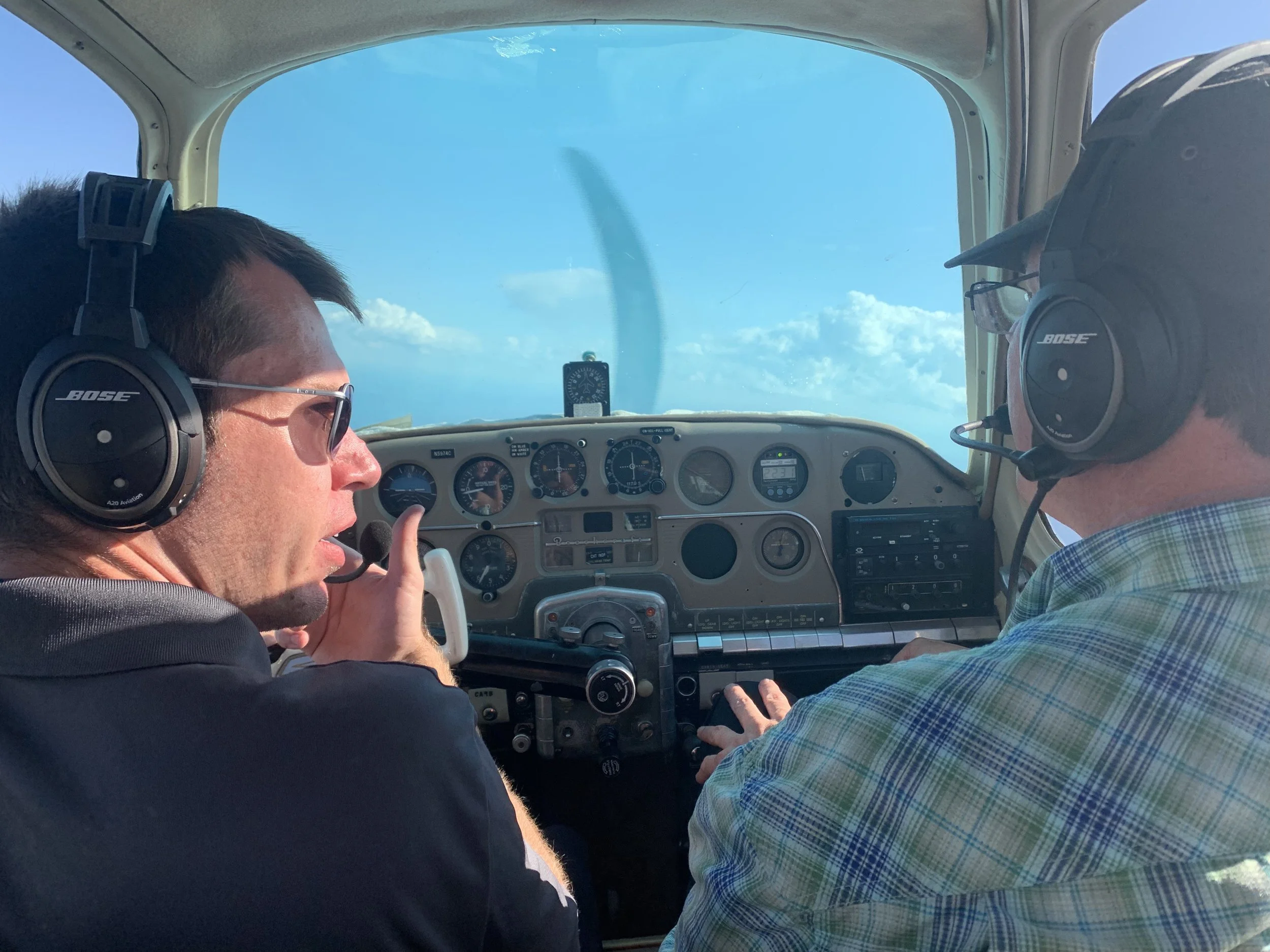 Two people wearing headsets in a small aircraft cockpit, focusing on flying controls and instruments, with a clear sky visible outside.