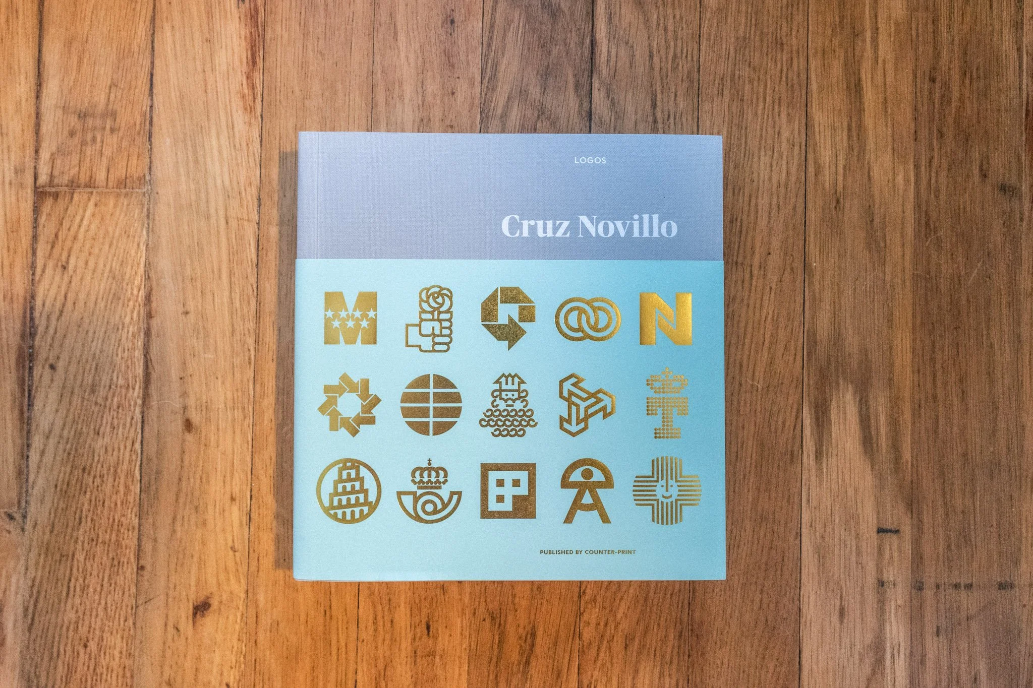 Cover of Cruz Novillo: Logos by Counter-Print placed on a wooden floor, featuring a grid of José María Cruz Novillo’s iconic logo designs stamped in gold foil.