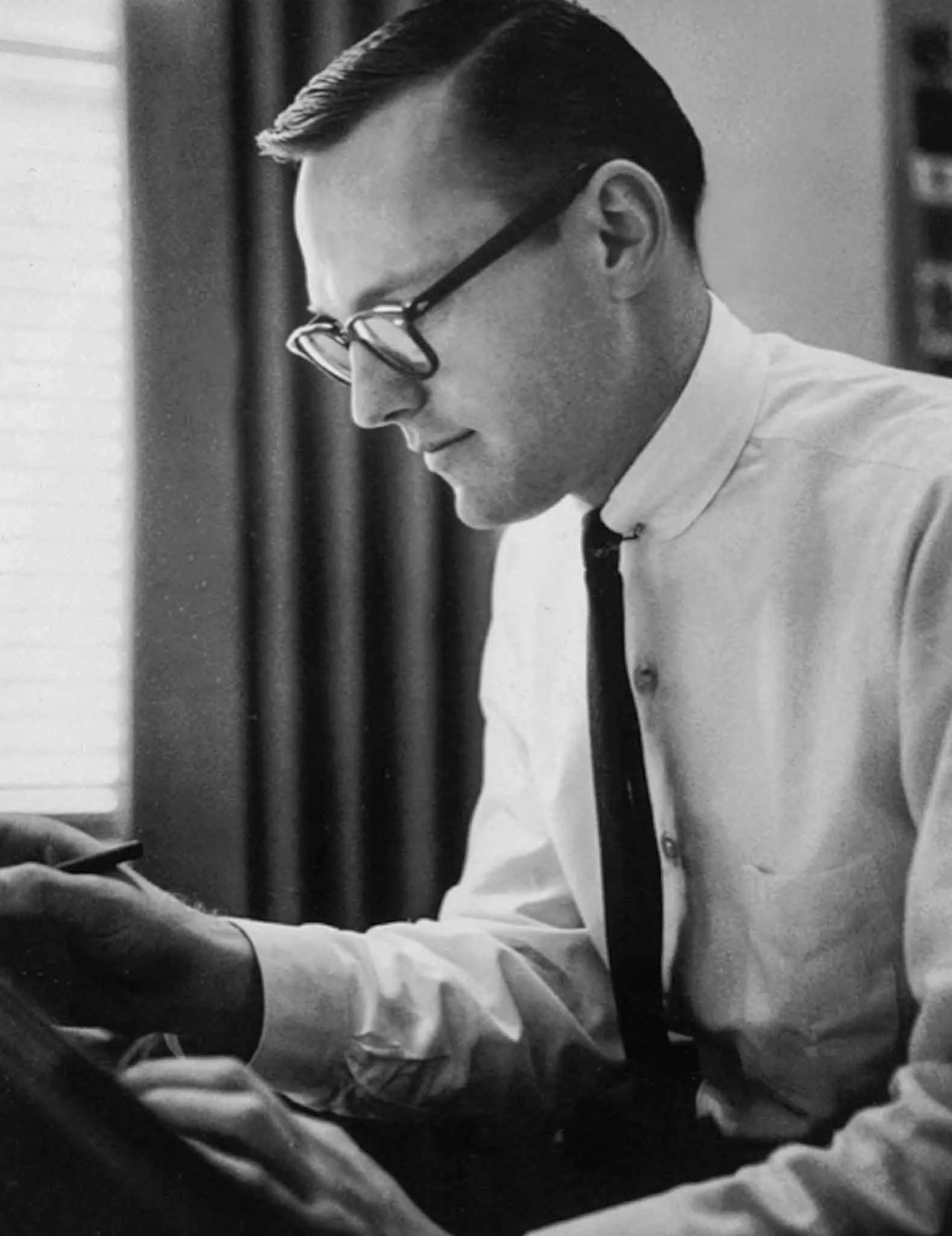 Black-and-white portrait of graphic designer Richard Danne seated at a drawing table, concentrating on his work while wearing a white dress shirt, narrow black tie, and thick-rimmed glasses.