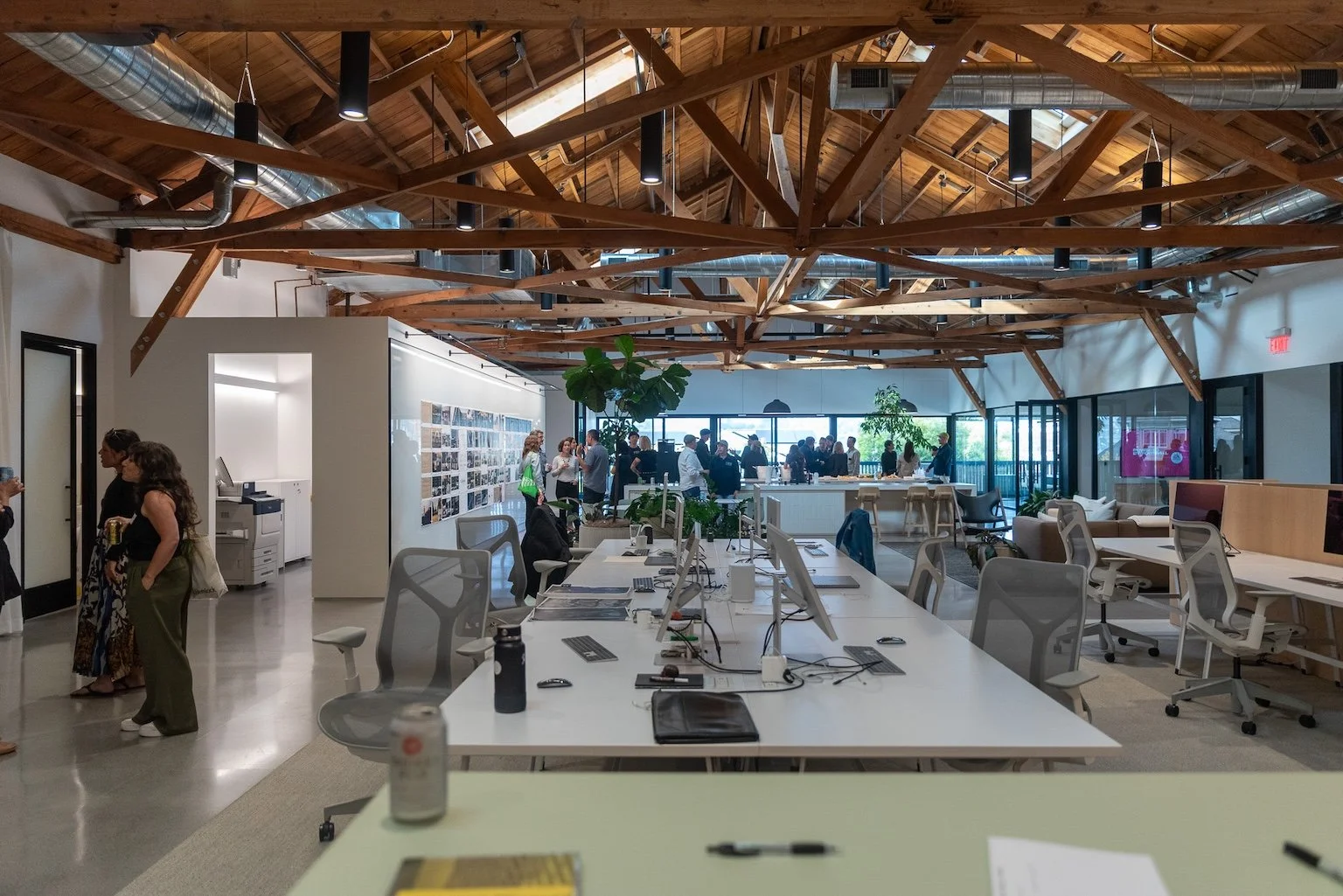 An expansive open-plan design studio featuring exposed wooden ceiling trusses, sleek white workstations, polished concrete floors, modern pendant lighting, and large windows with natural light.