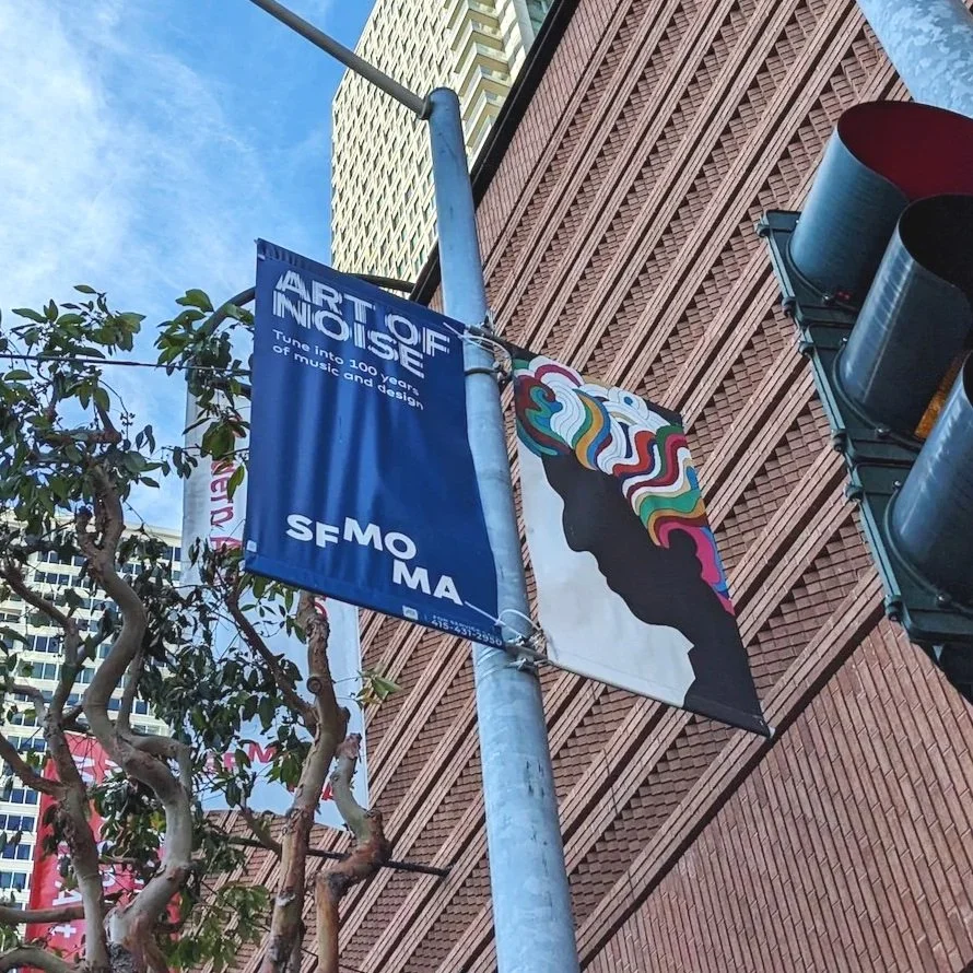 Street view of SFMOMA banners on a pole, one reading “Art of Noise” and another showing Milton Glaser’s Dylan profile artwork, with a traffic light and tall buildings in the background.