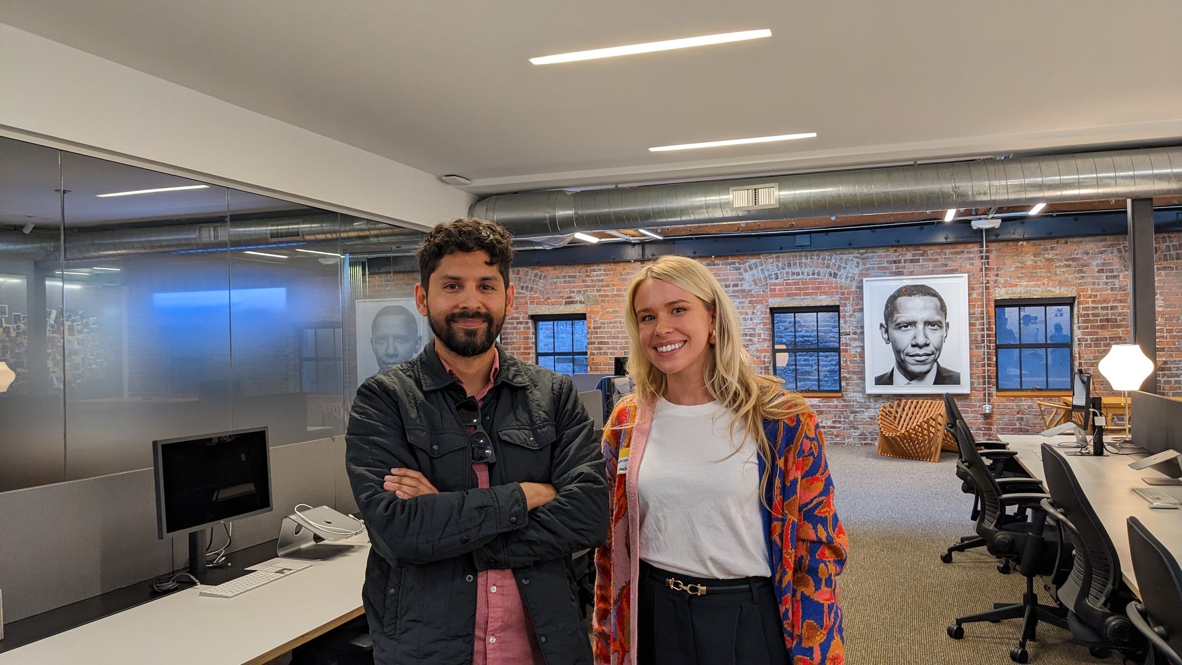 Two people standing and smiling inside an open-concept office with exposed brick walls, shared desks, and framed portrait photography in the background.