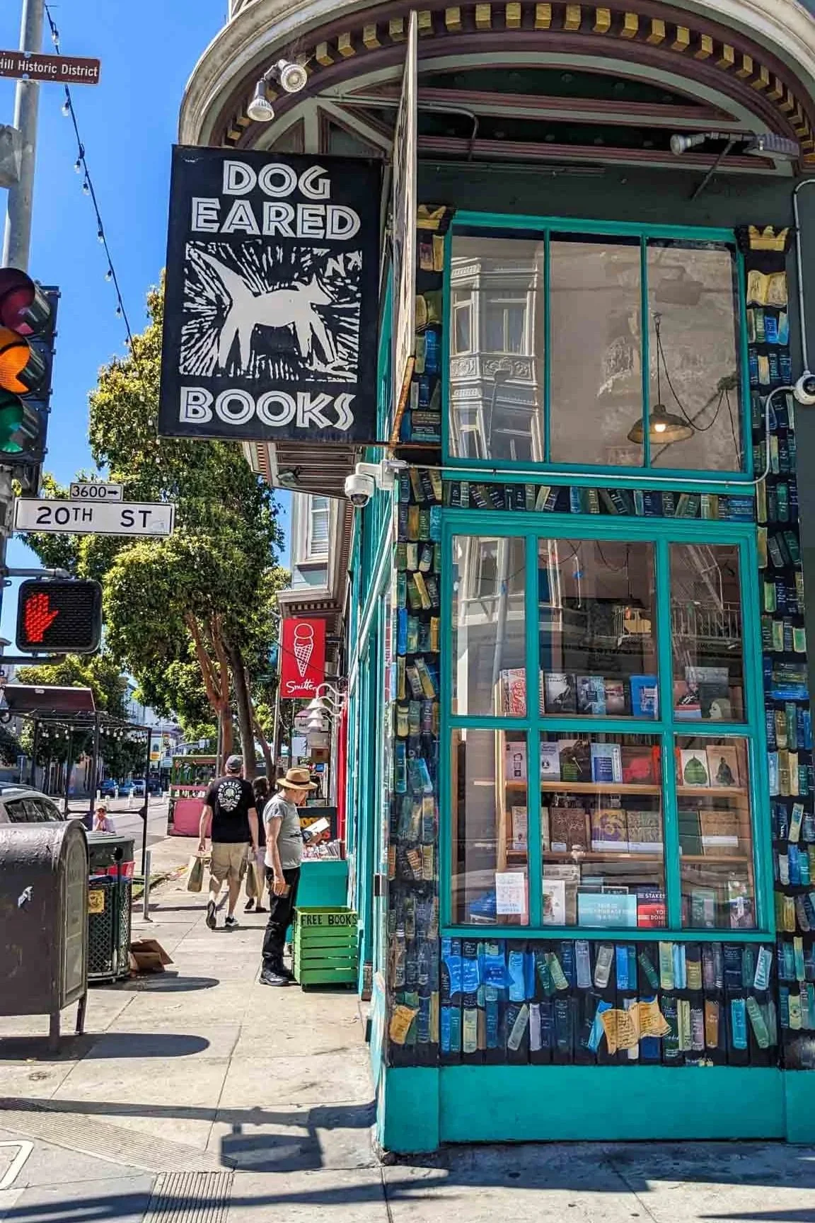 Exterior view of Dog Eared Books on the corner of 20th Street in San Francisco's Mission District. The storefront features bright turquoise trim, a large hanging bookstore sign, and windows filled with books on a sunny day.