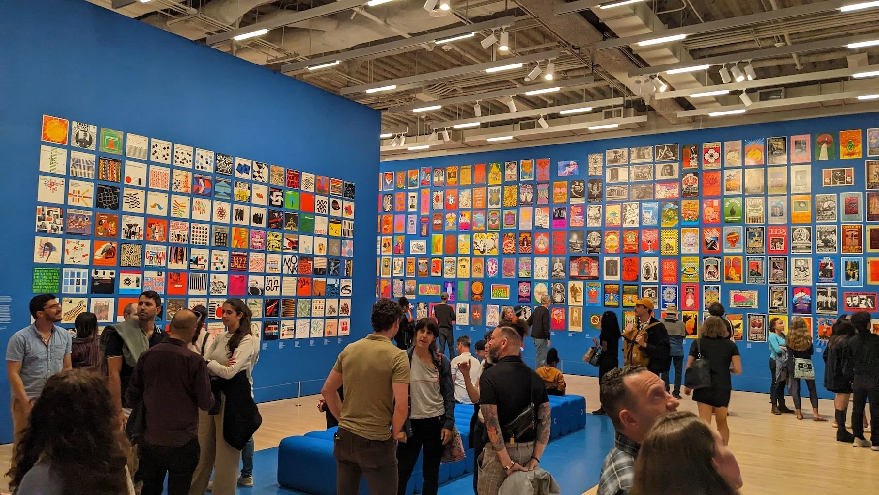 Visitors gather in a large gallery space with bright blue walls covered in a grid of album covers and posters at SFMOMA.