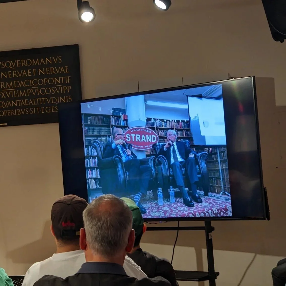 Audience members watch a presentation at Letterform Archive as a large screen displays Richard Danne and Michael Bierut in conversation during their interview at The Strand Book Store in New York City.