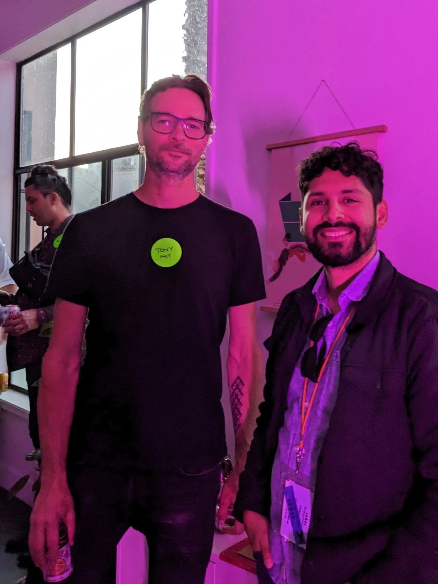 Two designers standing together inside Pact Studio during San Francisco Design Week, illuminated by pink lighting, with large Mission District windows behind them.