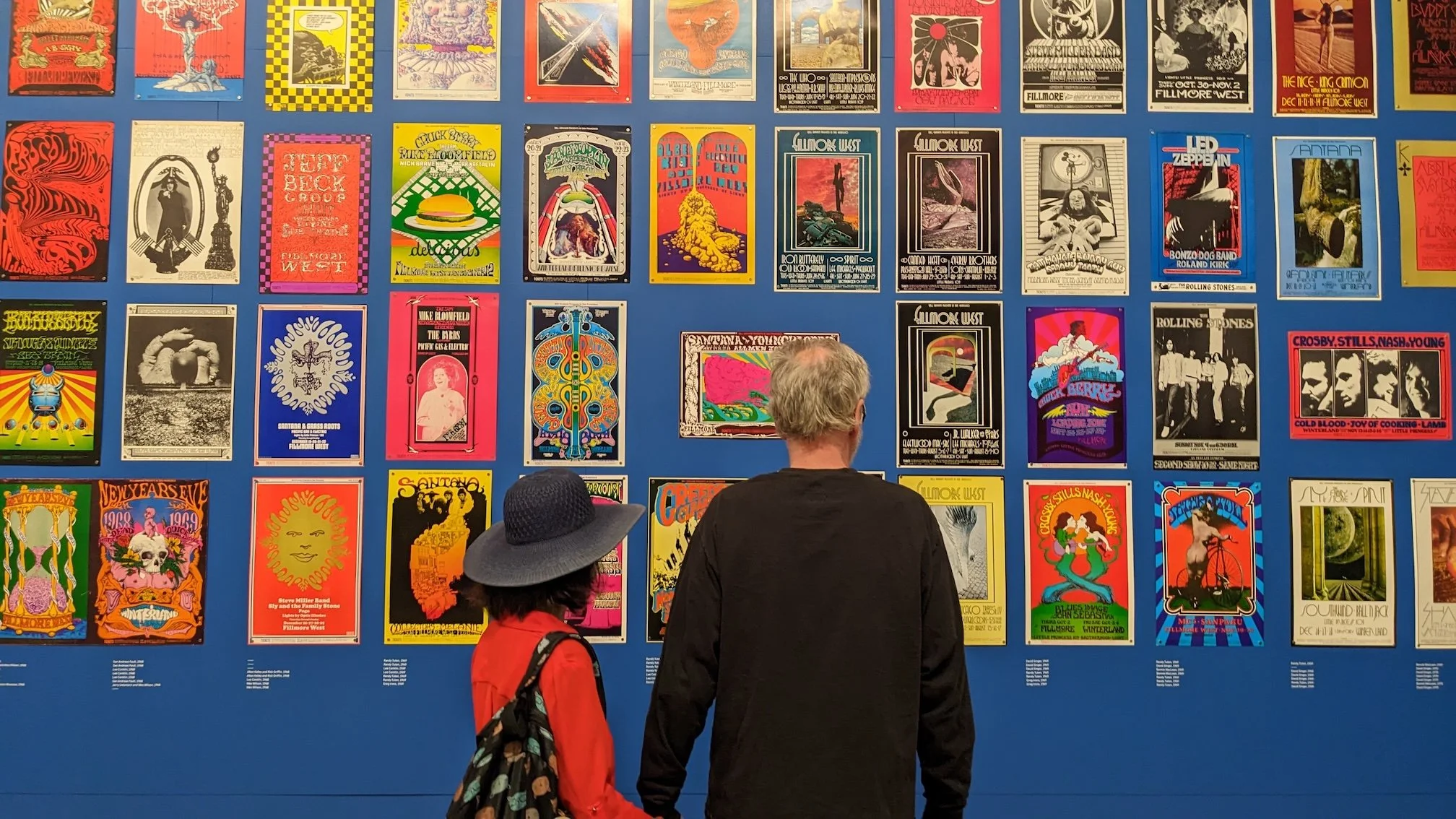 Two people stand facing a wall filled with colorful concert posters arranged in a grid on a bright blue background at a museum exhibition.
