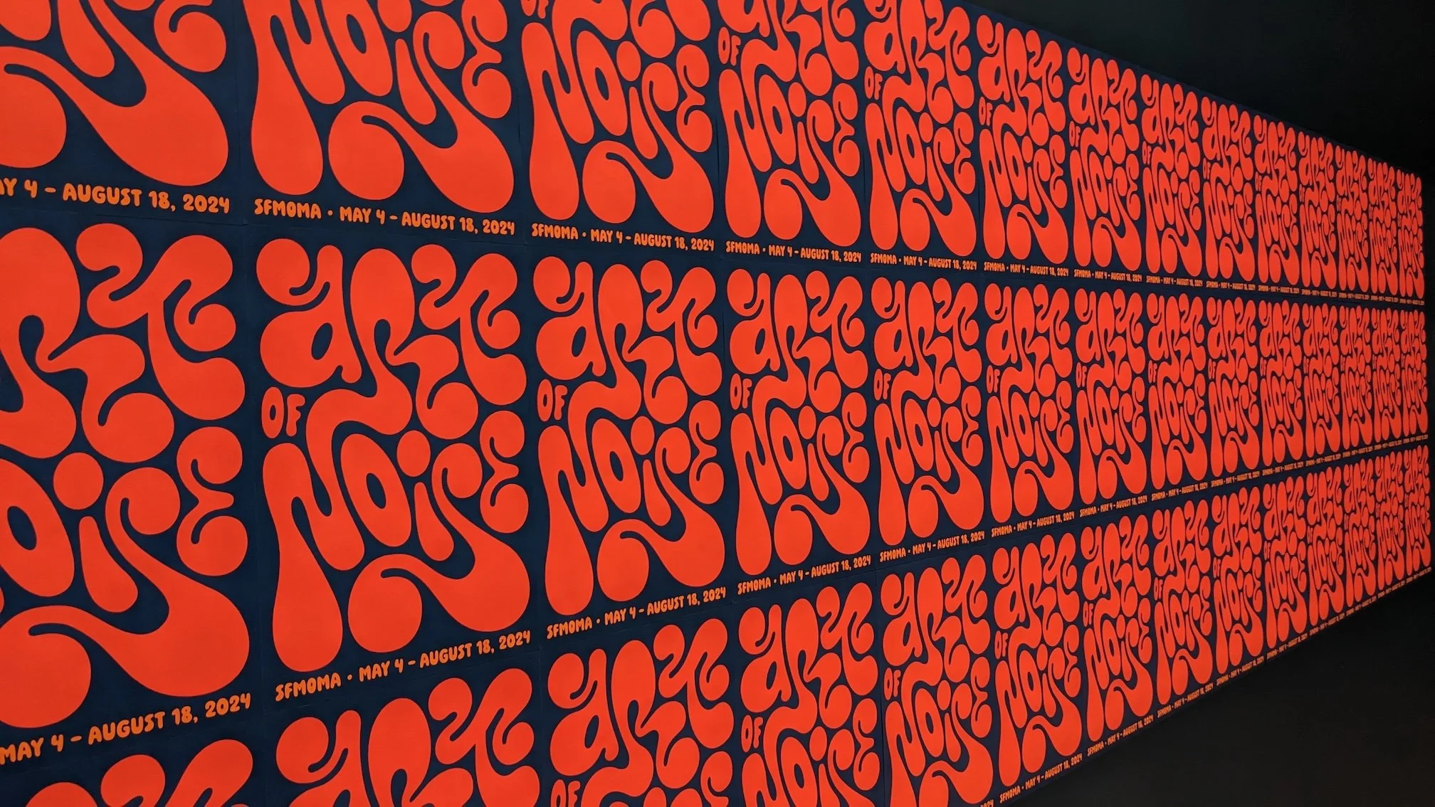 A wall covered in repeating red “Art of Noise” posters with bold, psychedelic lettering on a dark blue background, installed in a grid across a museum gallery.