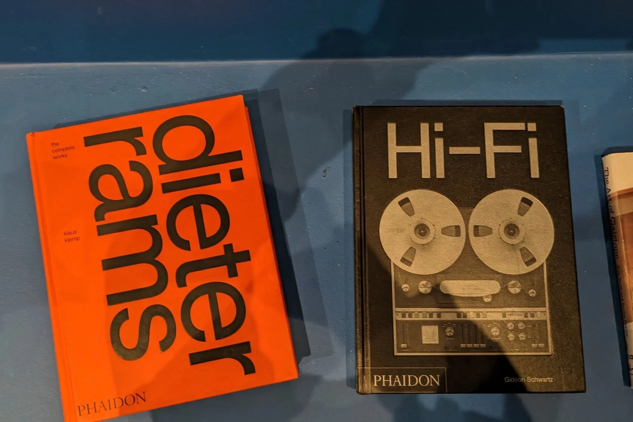 Two books displayed on a blue surface: a bright orange Dieter Rams: The Complete Works book with bold black typography and a black Hi-Fi book featuring a reel-to-reel tape recorder on the cover.