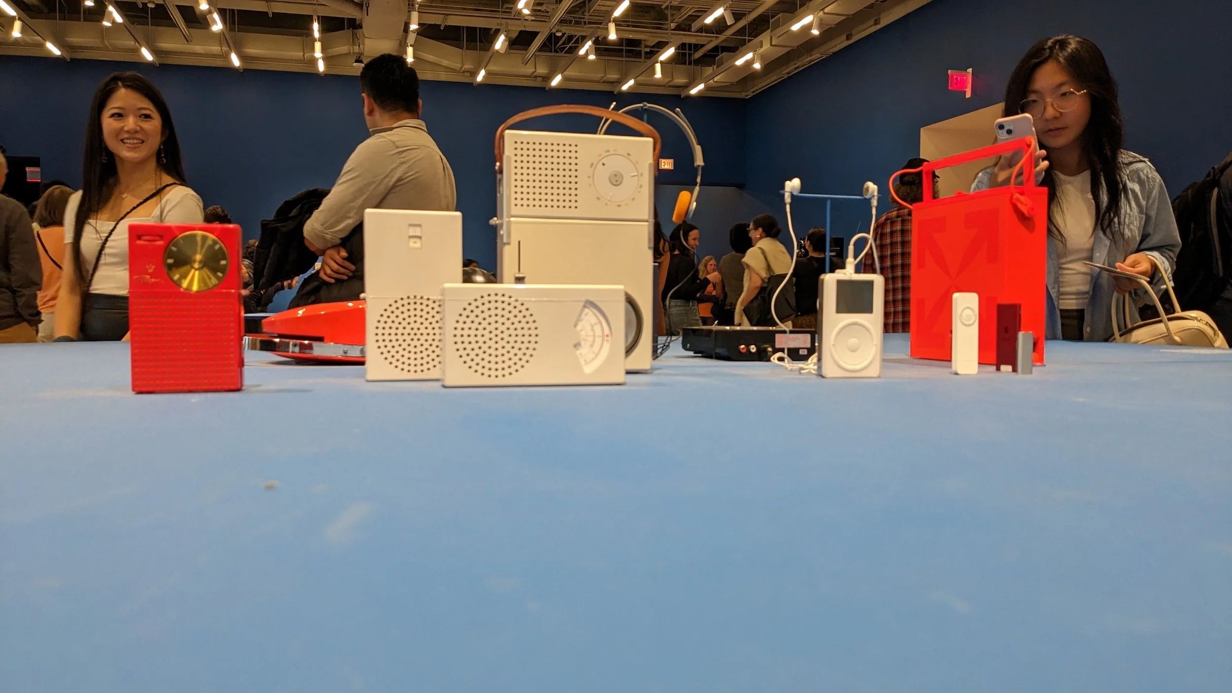 A museum display of vintage and modern audio devices, including colorful mid-century radios with knobs and dials alongside sleek white minimalist speakers, arranged on shelves against a white wall.