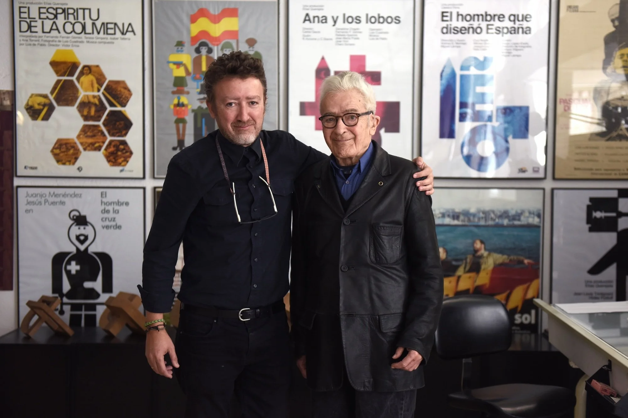 José María Cruz Novillo standing beside his son, Pepe Cruz Novillo Jr., in front of a wall of posters featuring work from across his design career.