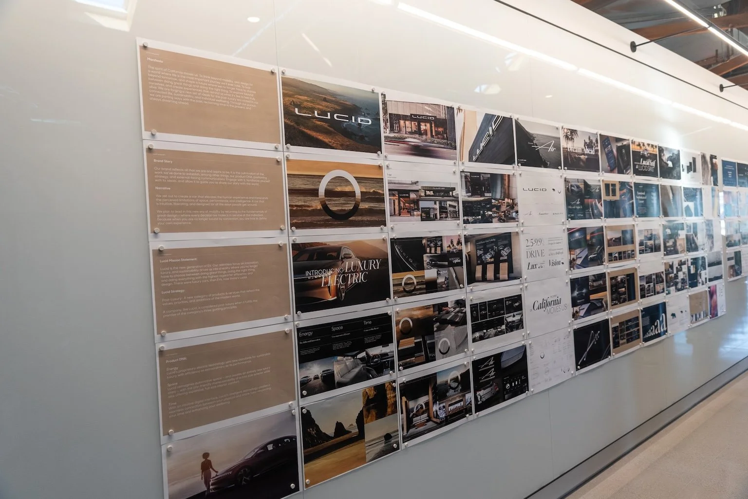 A long white studio wall filled with a clean grid of printed case studies and brand collateral for Lucid Motors, featuring typography, strategy text, and luxury electric vehicle photography held in place with white magnets