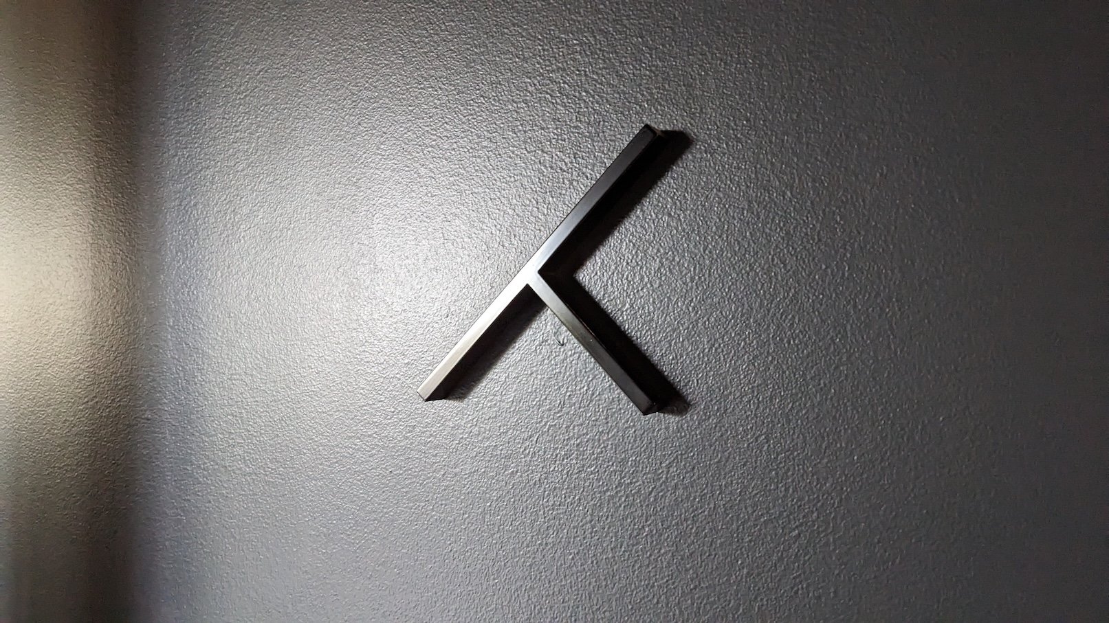 A minimal, black dimensional metal Tolleson logomark mounted on a textured grey studio wall under dramatic side directional lighting.