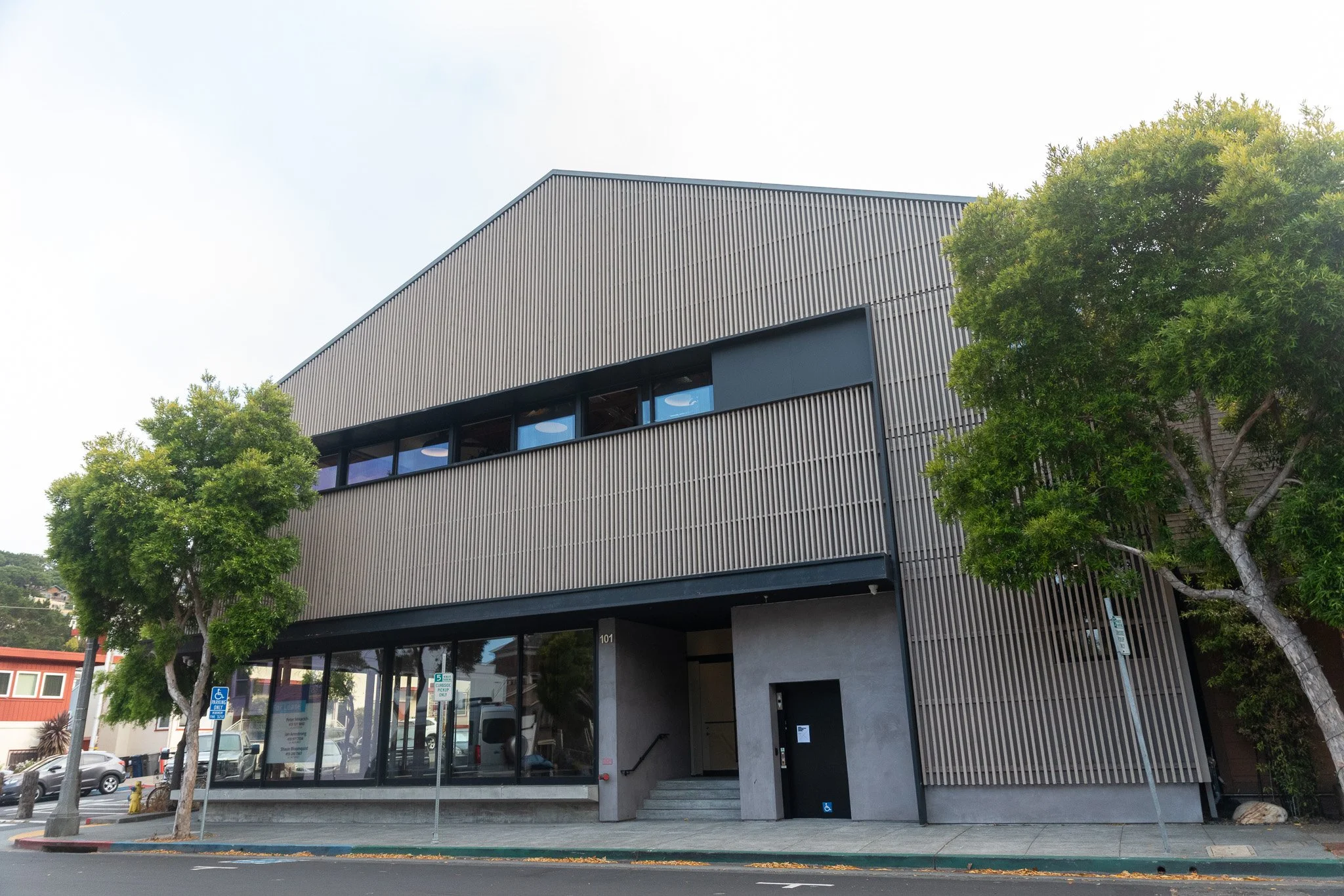 A modern two-story building in Sausalito featuring vertical wood slatted siding, a gabled roofline, ground-floor glass windows, concrete entryway steps, and surrounding leafy green trees.