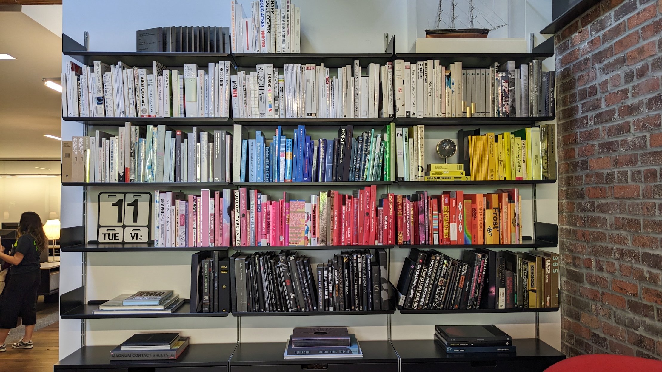 Color-coded bookshelf in the Godfrey Dadich Partners studio, arranged from white to black with rows of design books, art monographs, and creative references, next to an exposed brick wall.
