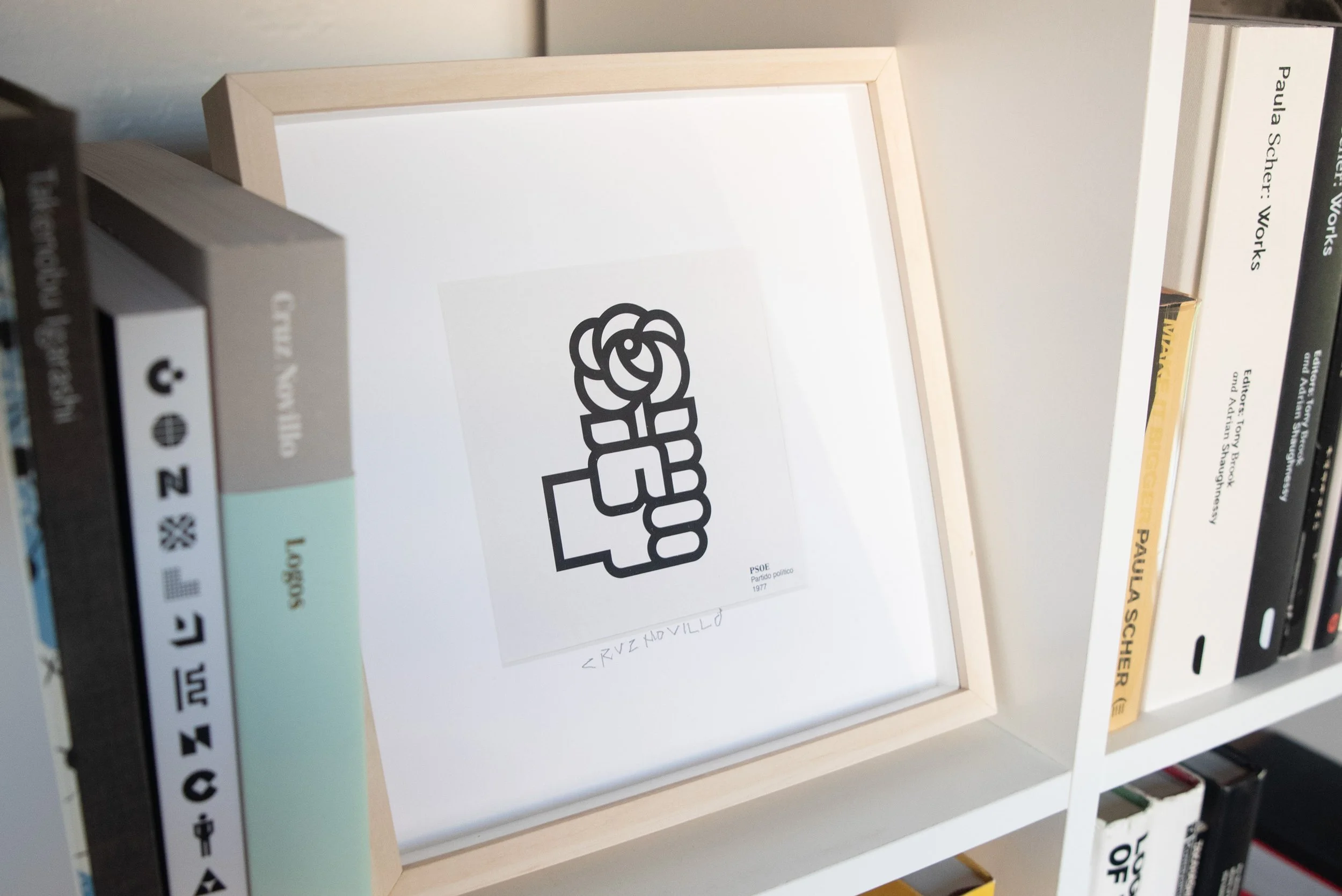 Framed print of José María Cruz Novillo’s 1977 PSOE fist-and-rose symbol displayed on a bookshelf beside several graphic design books, including Cruz Novillo: Logos by Counter-Print.