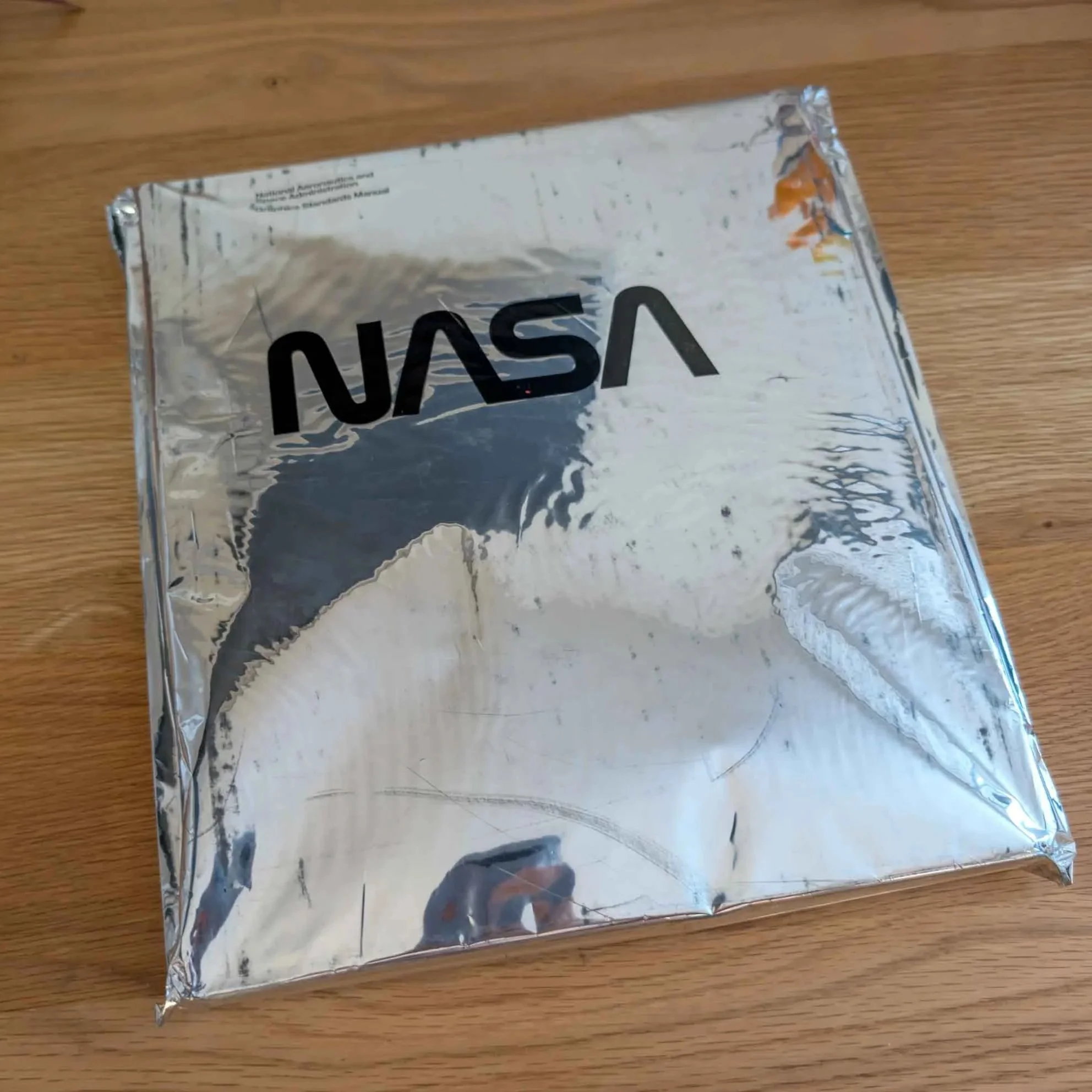 Silver-foil hardcover copy of NASA: National Aeronautics and Space Administration Graphics Standards Manual by Richard Danne and Bruce Blackburn, still sealed in its original protective shrink wrap, resting on a wooden tabletop.
