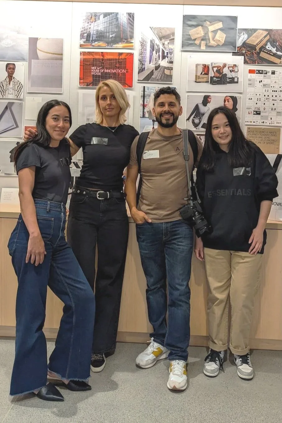 A group of four smiling people posing together in the studio, standing in front of a white wall covered with pinned Nike branding photos, retail mockups, and case study prints.