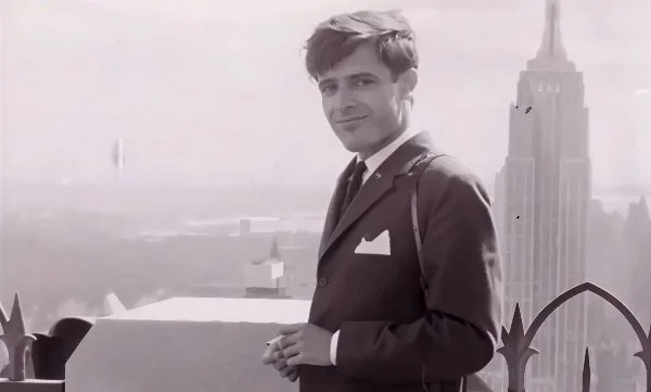 Black-and-white portrait of a young José María Cruz Novillo in a suit, standing on a rooftop terrace with the New York City skyline and the Empire State Building in the background.