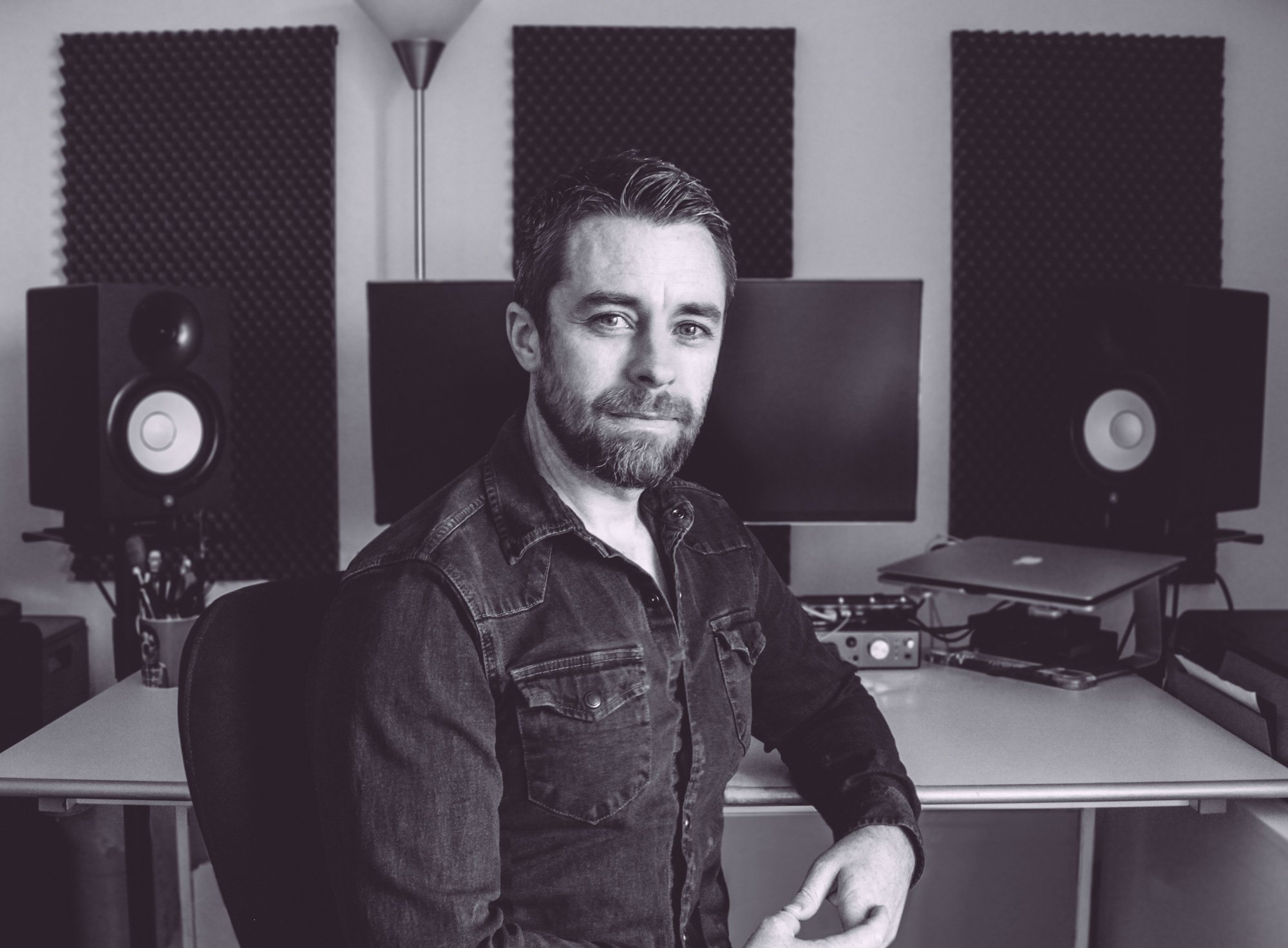 Gary Kelly. A graphic designer and music producer sitting in a music studio with soundproofing foam on the walls, two large speakers, a computer, and recording equipment.
