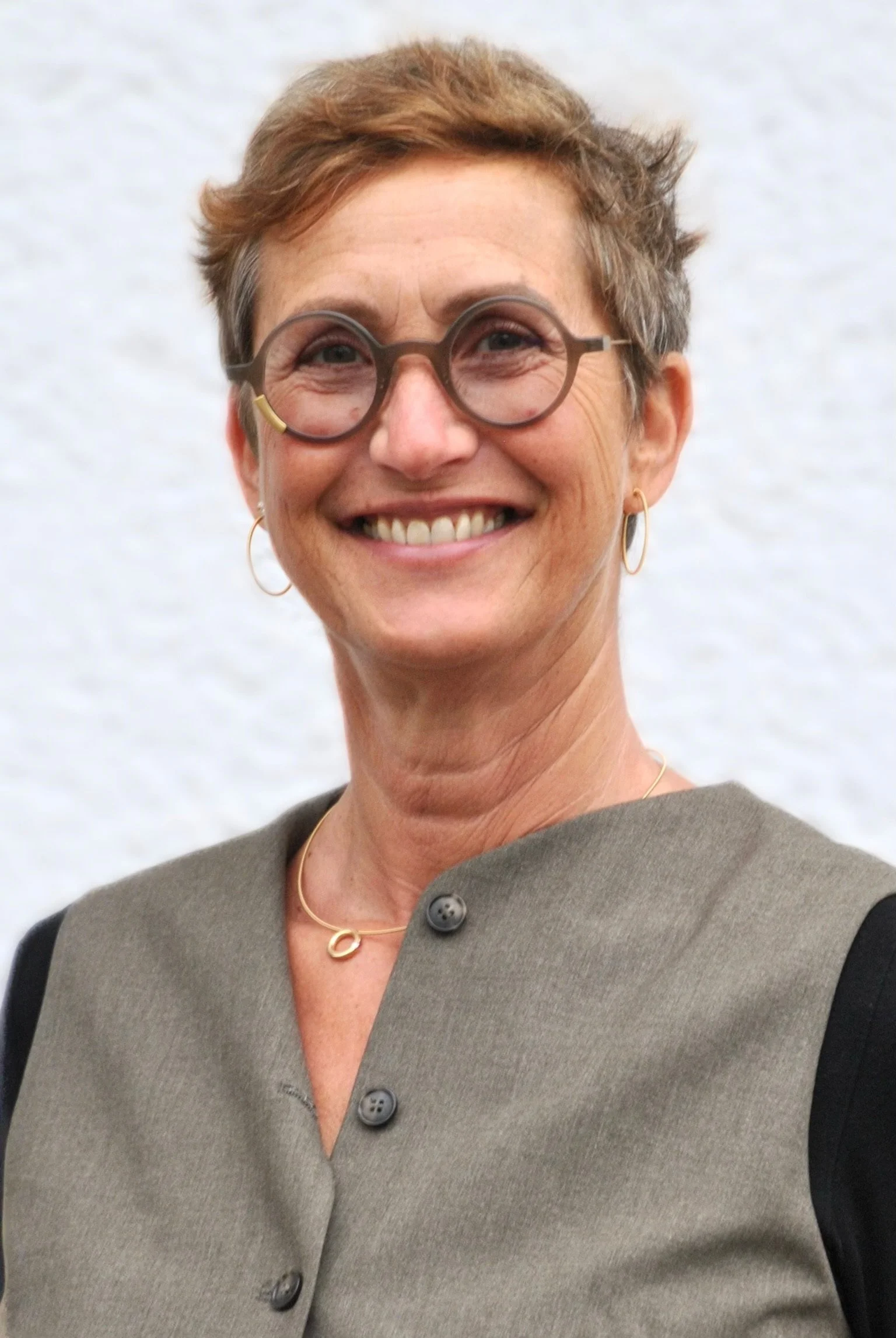 Nahaufnahme einer lächelnden Frau mit kurzen, lockigen braunen Haaren und eleganten runden Brillengläsern, trägt ein bunt gemustertes Hemd und eine zarte Goldkette mit Anhänger.