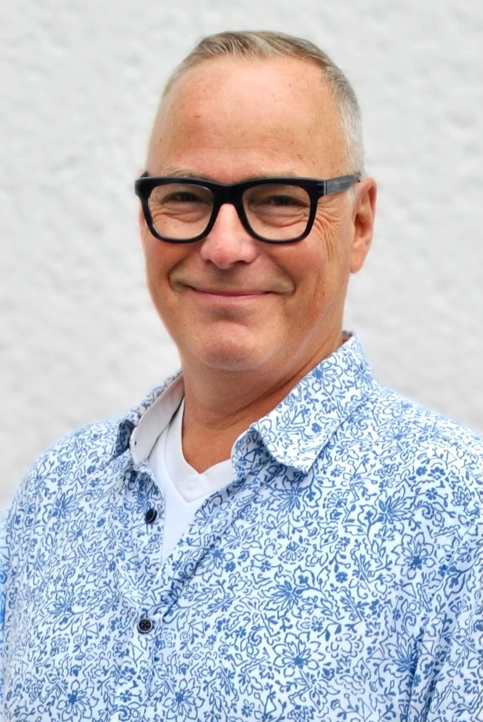 Nahaufnahme eines lachenden Mannes mit Brille und blauer Hemd vor weißer Wand.