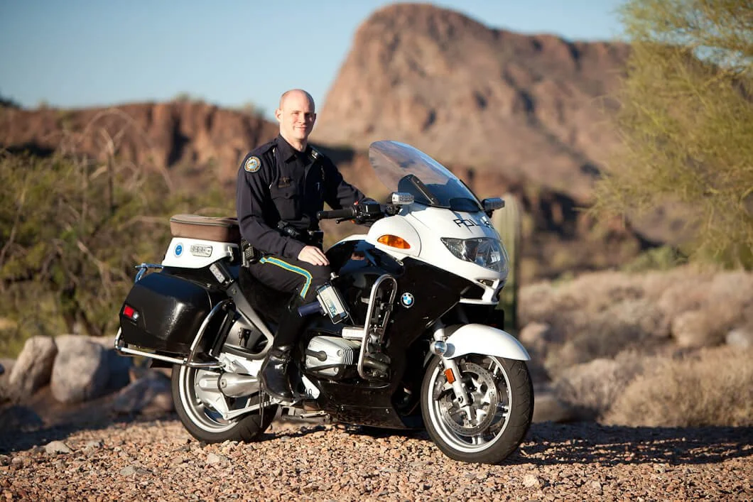 A police officer in uniform sitting on a black and white police motorcycle in a desert landscape with mountains and sparse vegetation in the background.