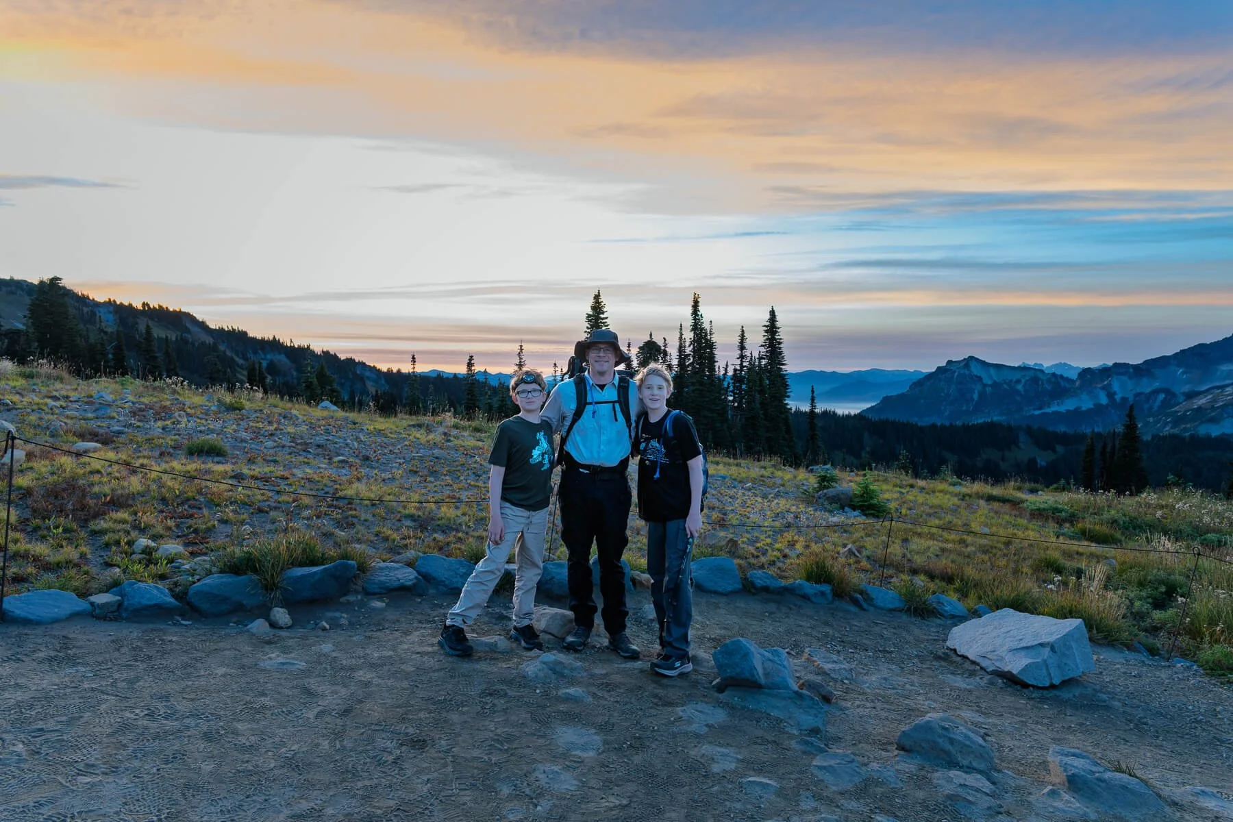 Three people standing together on a rocky trail during sunset with mountains and trees in the background.