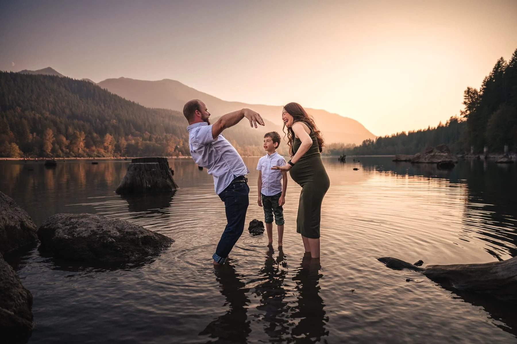 A family of three holding hands in a river at sunset, with mountains and trees in the background.