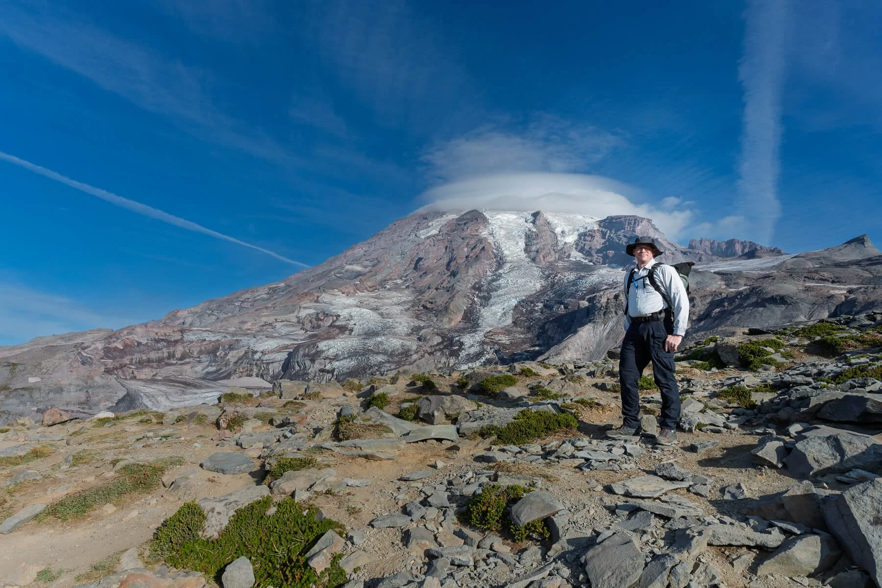 Hiker standing on rocky terrain with a mountain and volcanic eruption in the background, under a blue sky with clouds and contrails.