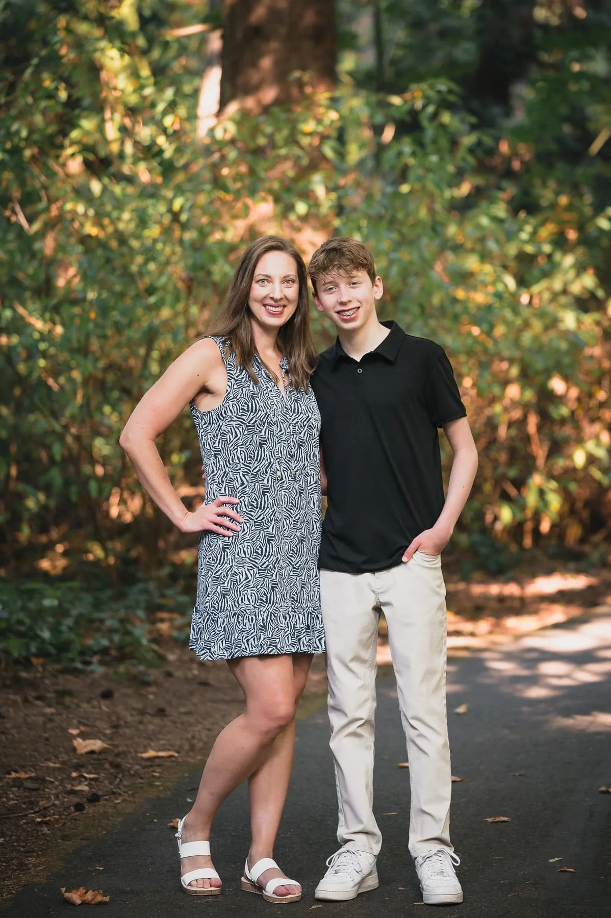 A smiling woman and a man standing outdoors in autumn with fallen leaves and colorful trees in the background.