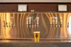 A glass of beer with the logo of Two Docs Brewing Company, placed on a bar counter with a blurred bar backdrop featuring multiple beer taps.