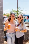 Two young women smiling and holding drinks at an outdoor event on a sunny day, with tents and people in the background.