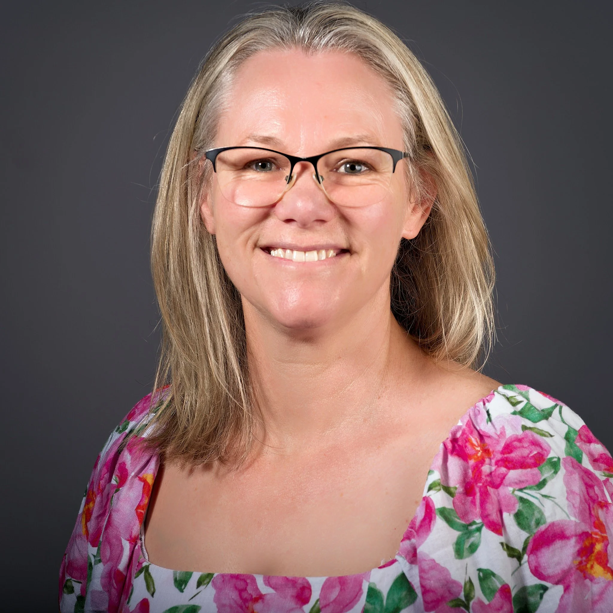 A woman with blonde hair, blue eyes, glasses, and a big smile wearing a pink, green, and white floral dress against a dark gray background.
