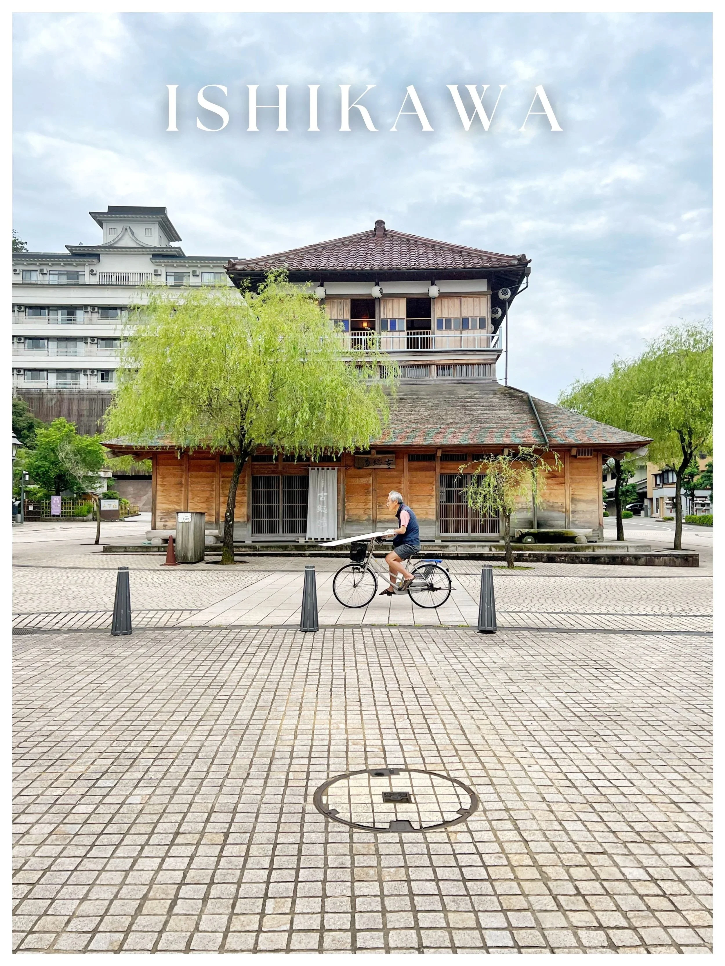 A man riding a bicycle past a traditional Japanese wooden building with a tiled roof, trees, and a cloudy sky.