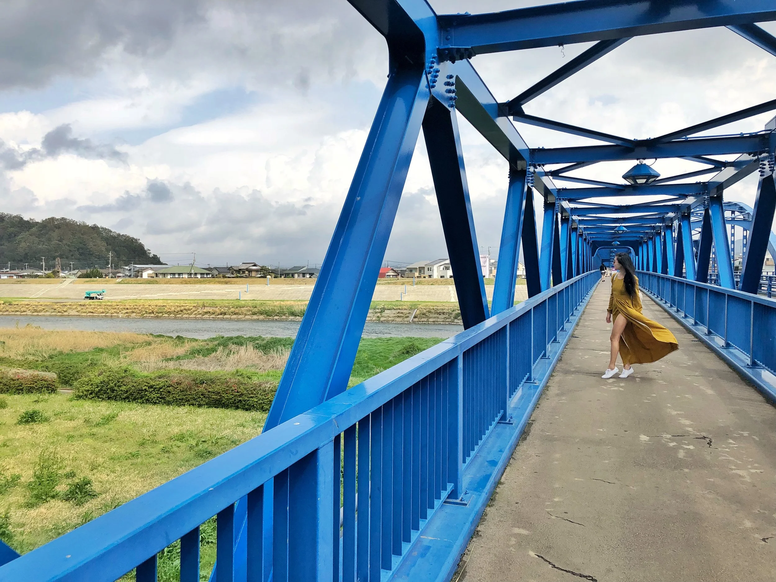 an asian woman with long black hair in a yellow dress walking on a blue bridge in the Japanese countryside on the way to check in for an onsen ryokan stay