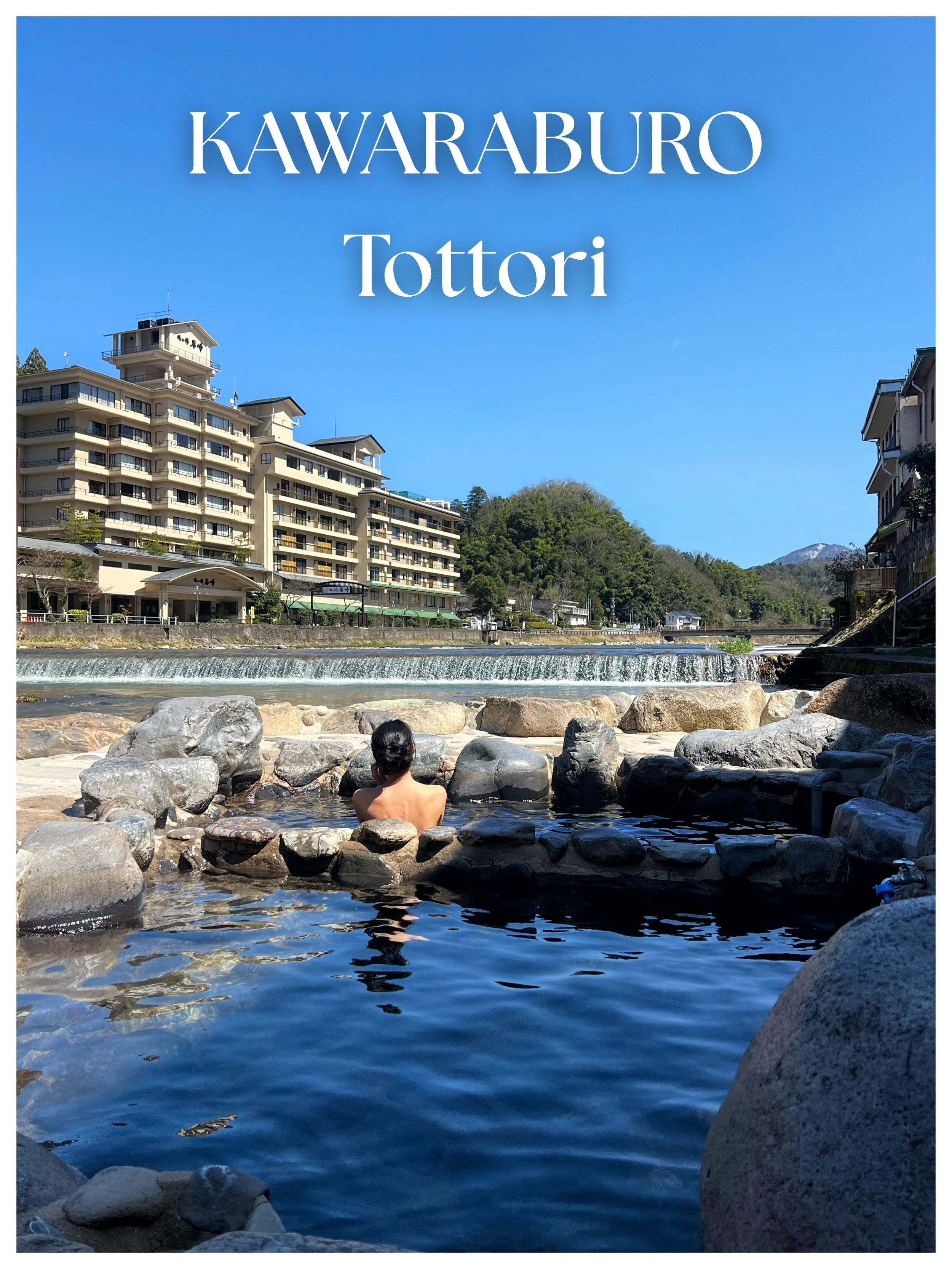 A person relaxing in a hot spring pool surrounded by large rocks, with a river and buildings in the background, under a clear blue sky in Togakushi, Japan.