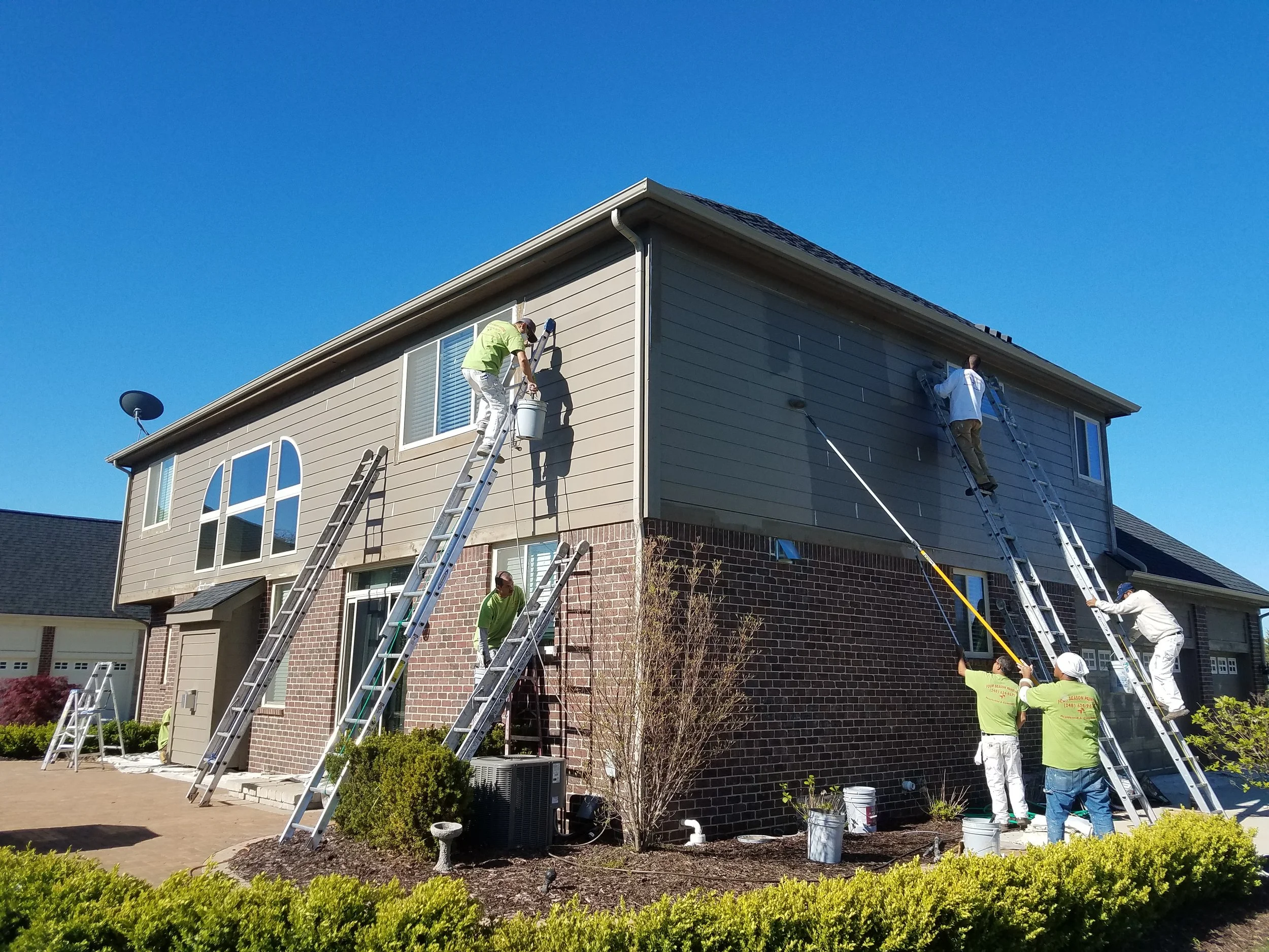 People painting the exterior of a two-story house with ladders on a sunny day.