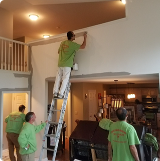 Three people wearing green shirts working on painting a wall inside a house, with one person on a ladder painting the upper part.