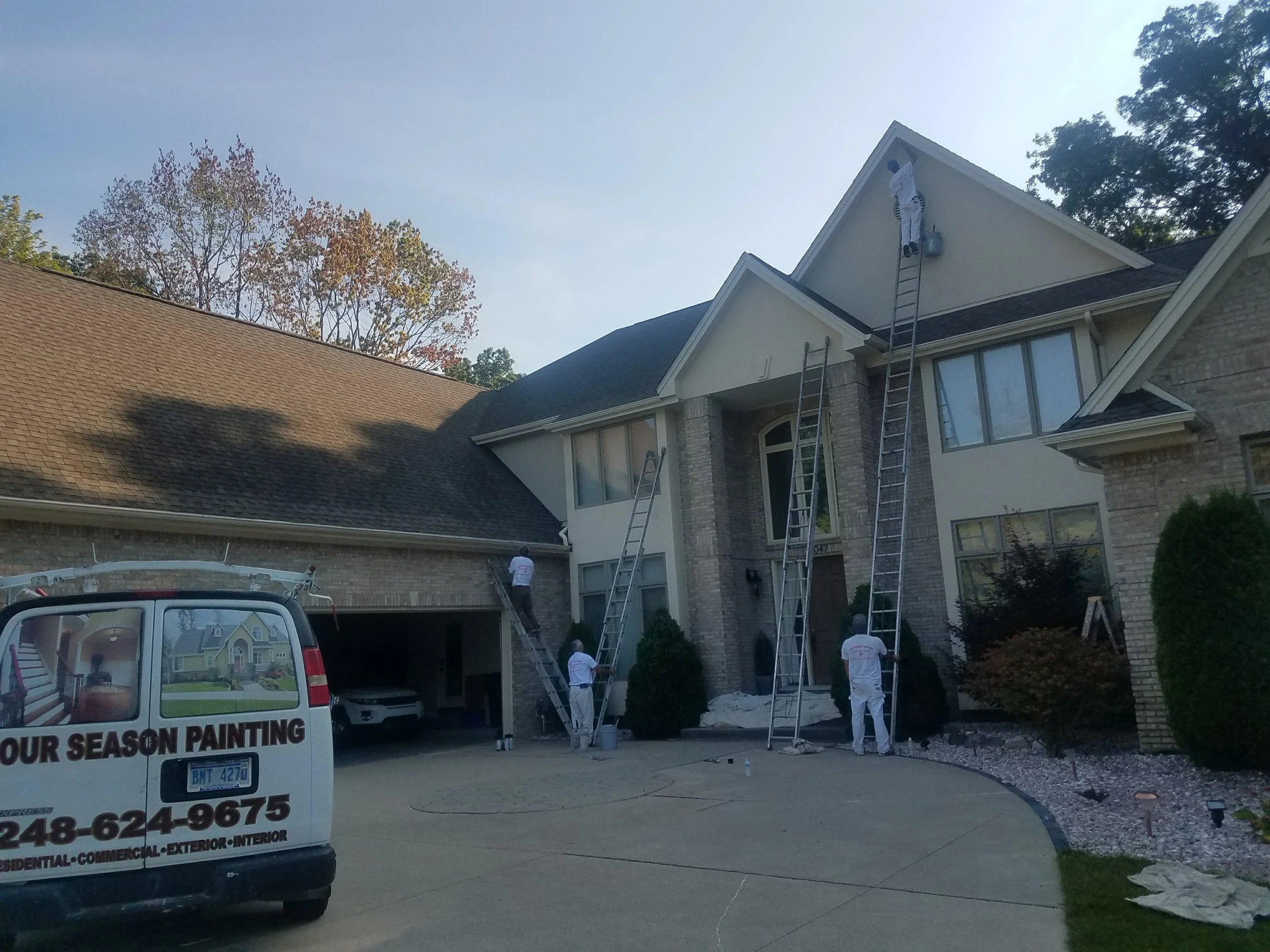 Workers painting the exterior of a large residential house with ladders, a van parked nearby, and trees in the background.