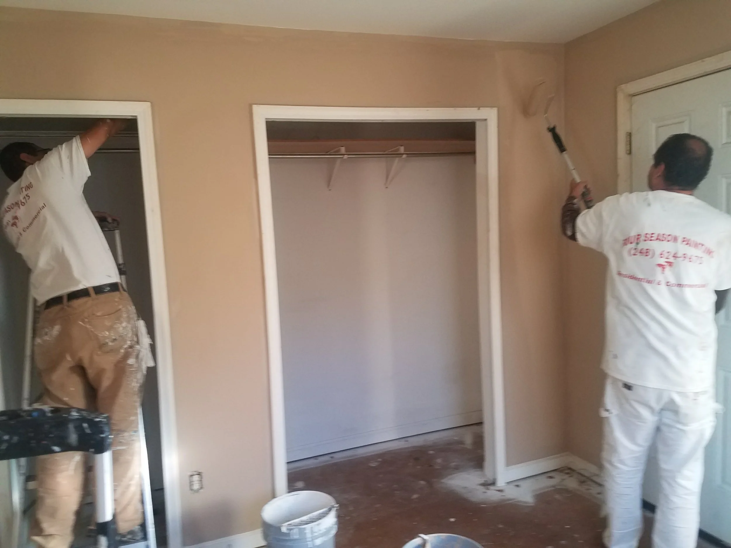 Two workers painting the wall of a room with a paint roller, with a closet and a door nearby.