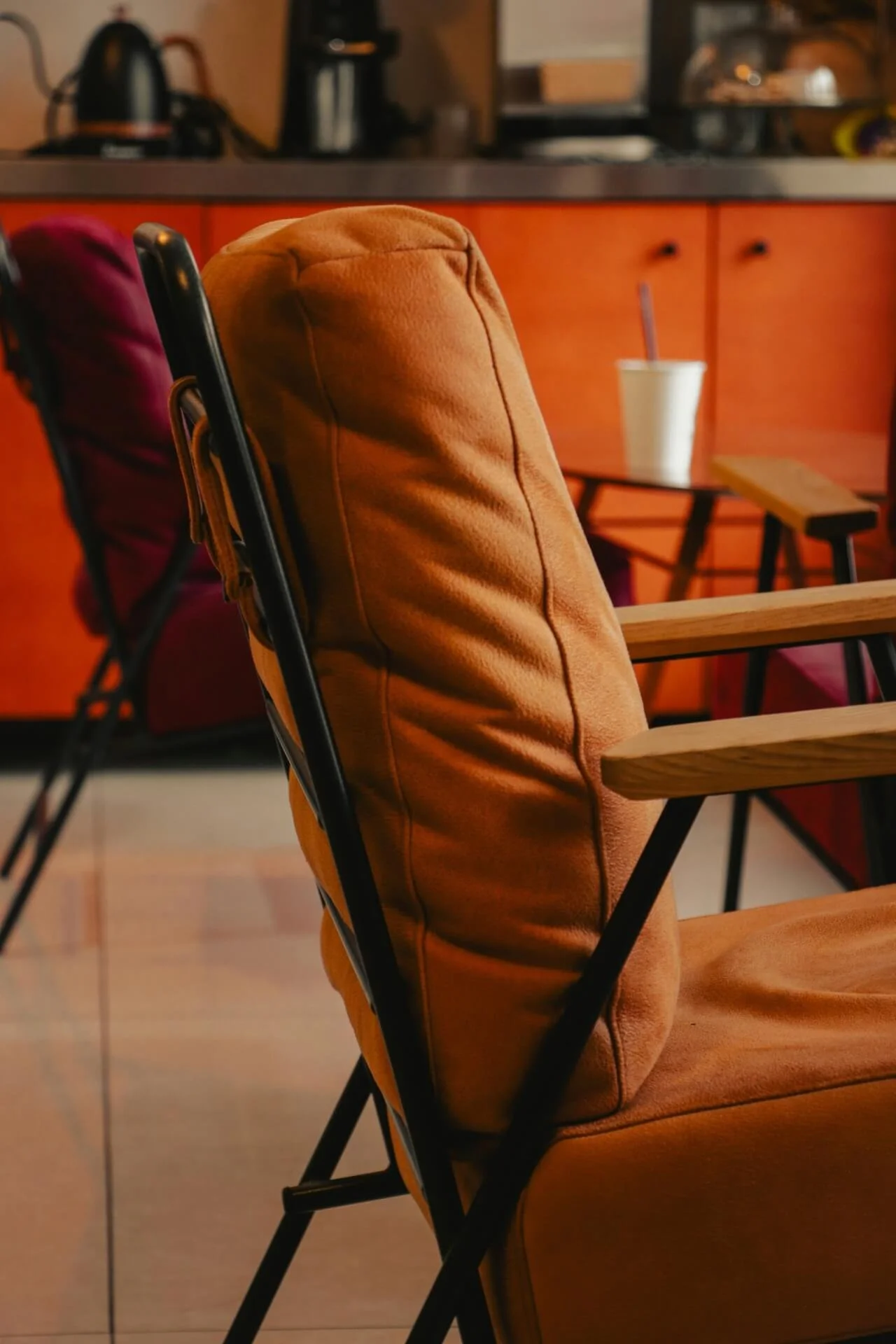 Close-up of a tan upholstered chair with black metal frame and wooden armrests in a cafe or restaurant setting.